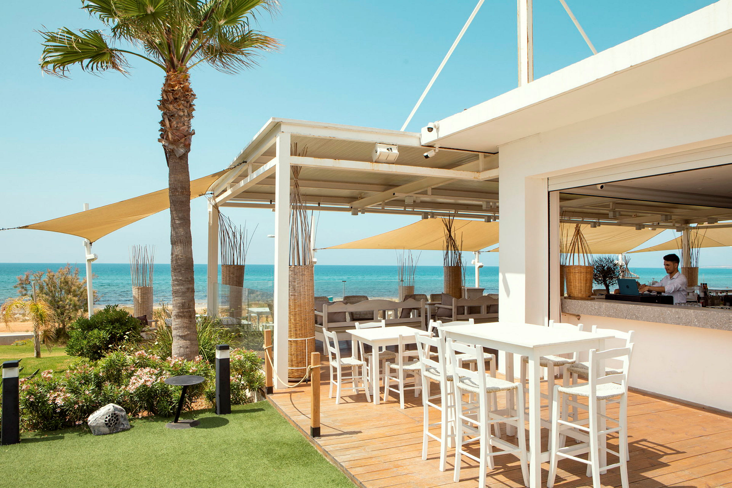 En utendørsrestaurant med bord og stoler, utsikt over havet og klar blå himmel. (AI-generert tekst)