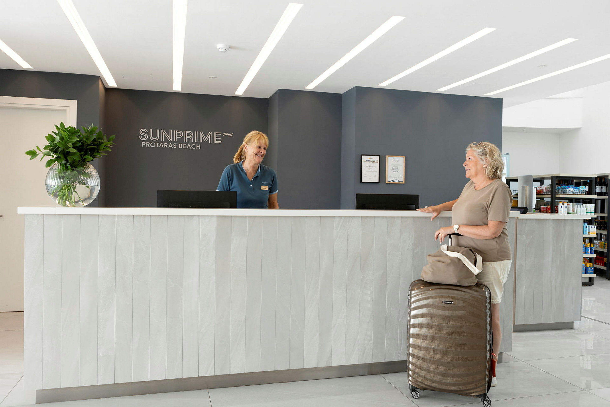 Resepsjonen på Sunprime Protaras Beach-hotellet med moderne innredning, inkludert en disk med en koffert og en håndveske plassert foran. (AI-generert tekst)