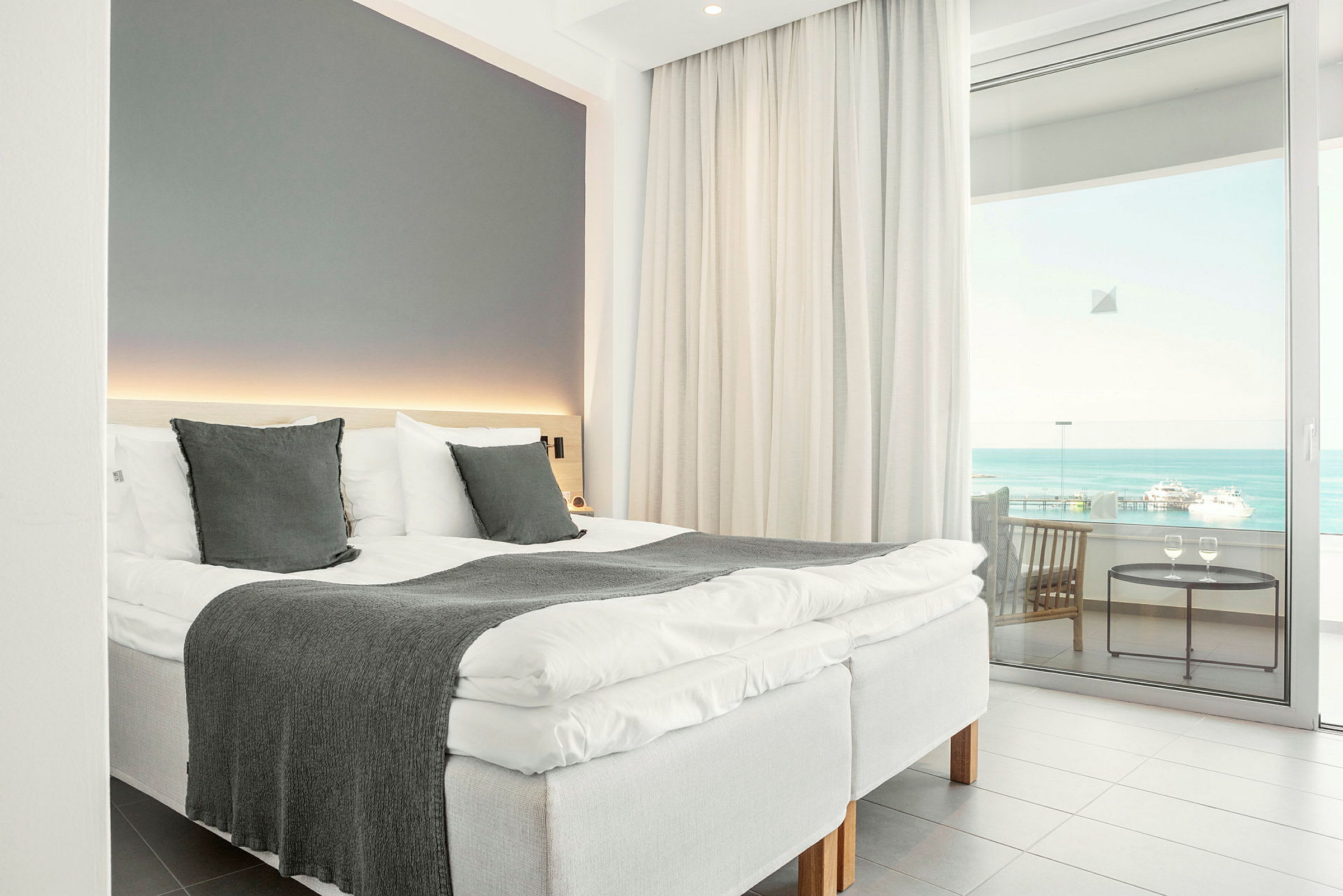 Et hotellrom med en oppredd seng, store vinduer og utsikt over havet. (AI-generert tekst)