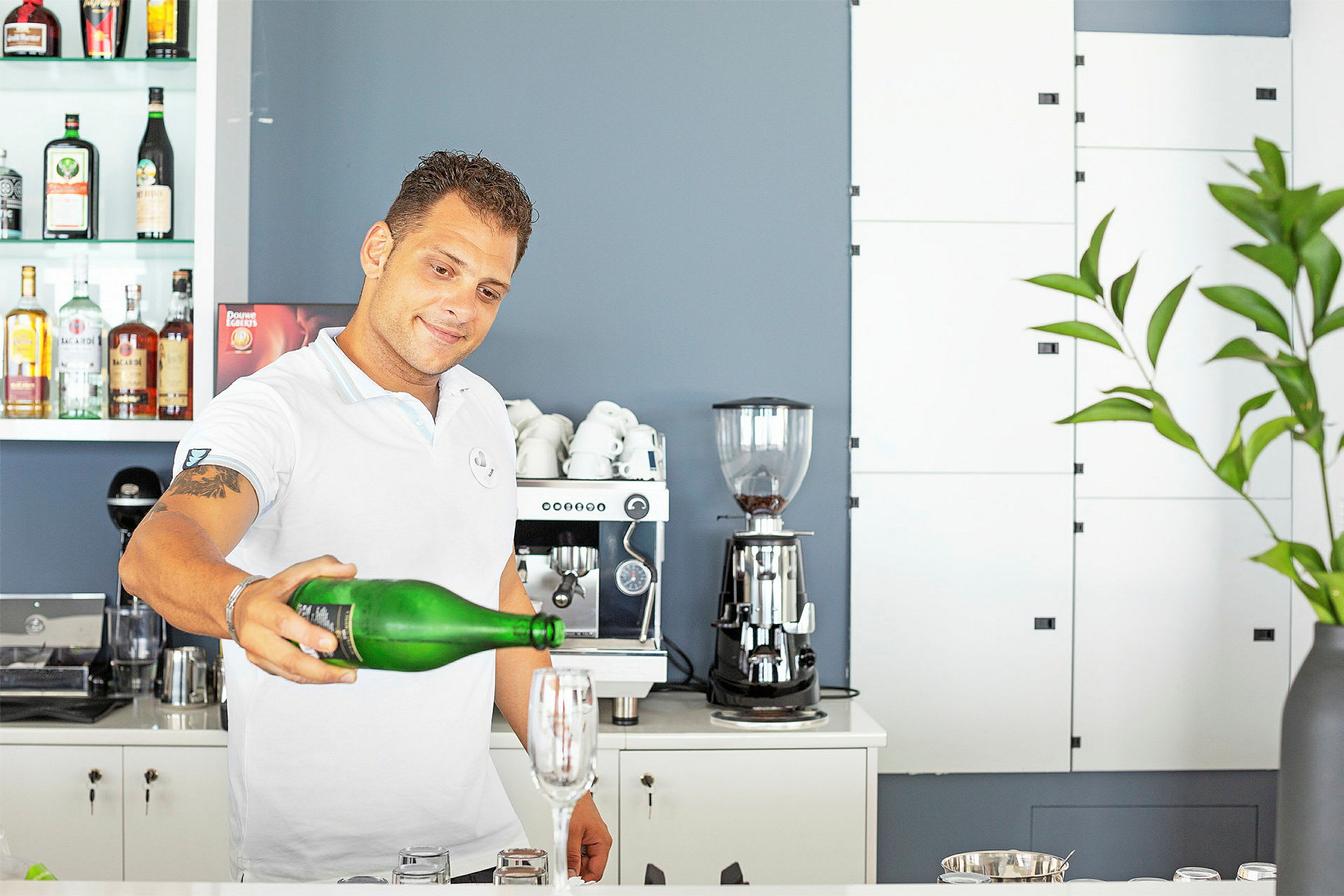 En person holder en grønn flaske og heller væske i et glass på en bardisk, omgitt av en kaffemaskin og flasker. (AI-generert tekst)