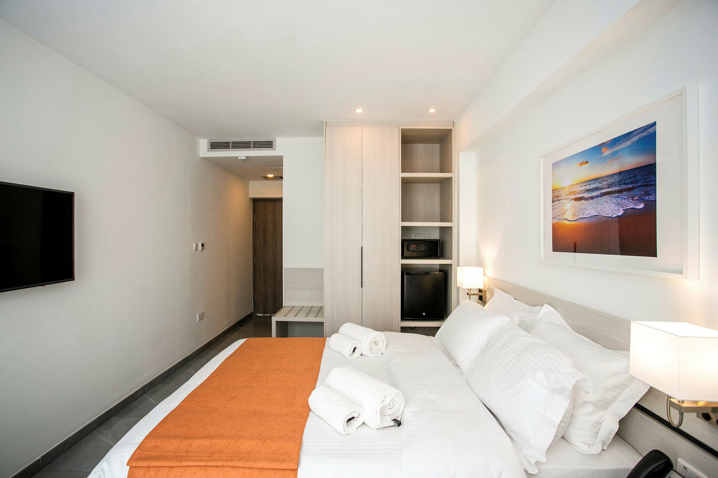 Et moderne hotellrom med en stor seng, to håndklær oppå, et skap, en TV på veggen og et maleri med motiv av en strand. (AI-generert tekst)