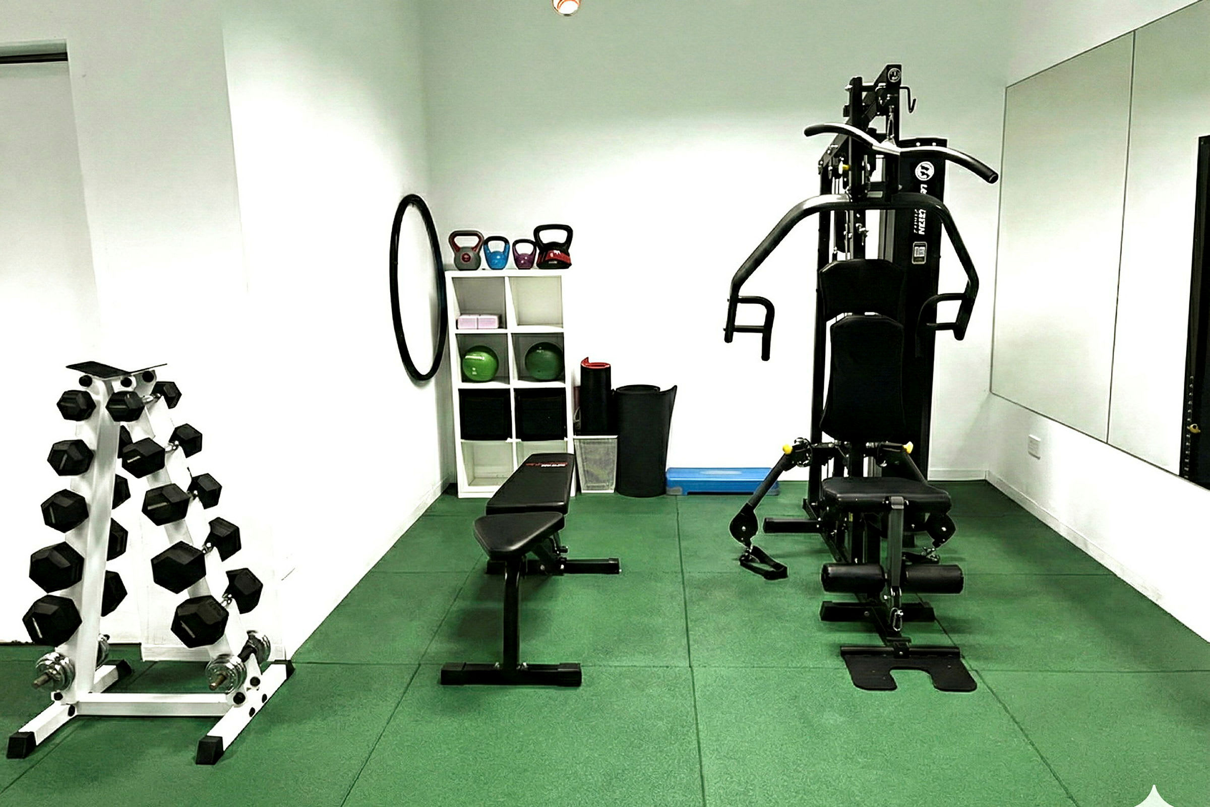 Et treningsstudio med treningsutstyr som manualer, benk, kettlebells og en treningsmaskin. Rommet har grønne gulvfliser og hvite vegger. (AI-generert tekst)