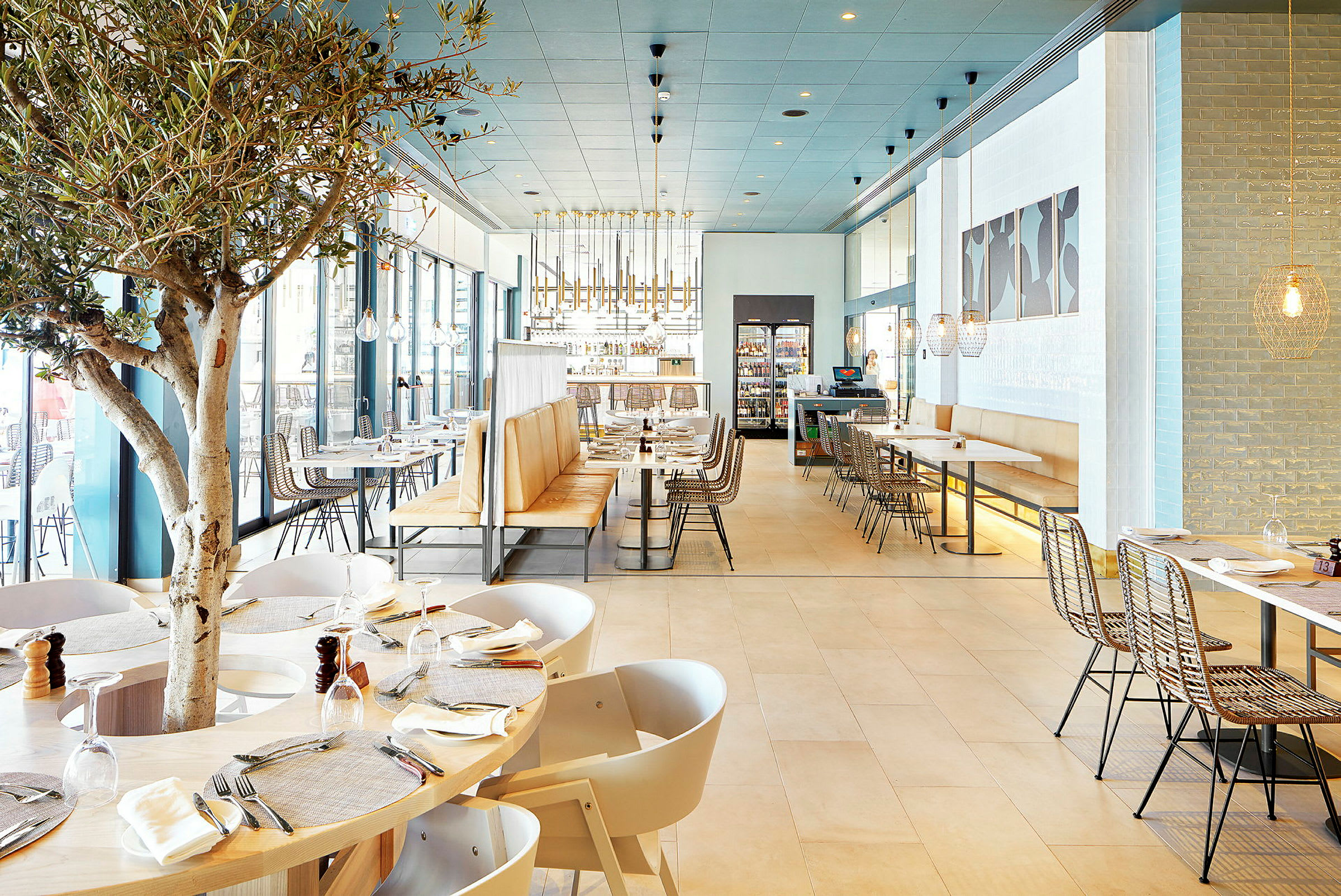Interiør av en moderne restaurant med tremøbler, lyse farger og et tre midt blant bordene. (AI-generert tekst)