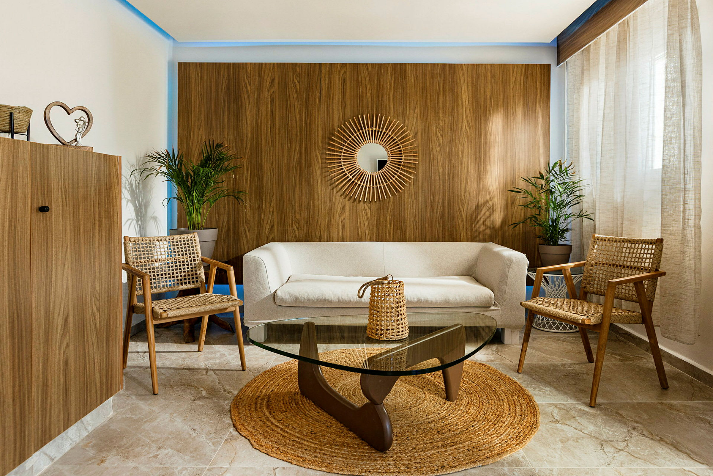 En stue med beige sofa, to flettede stoler, et glassbord og grønne planter. Et rundt speil henger på en trefinérvegg. (AI-generert tekst)