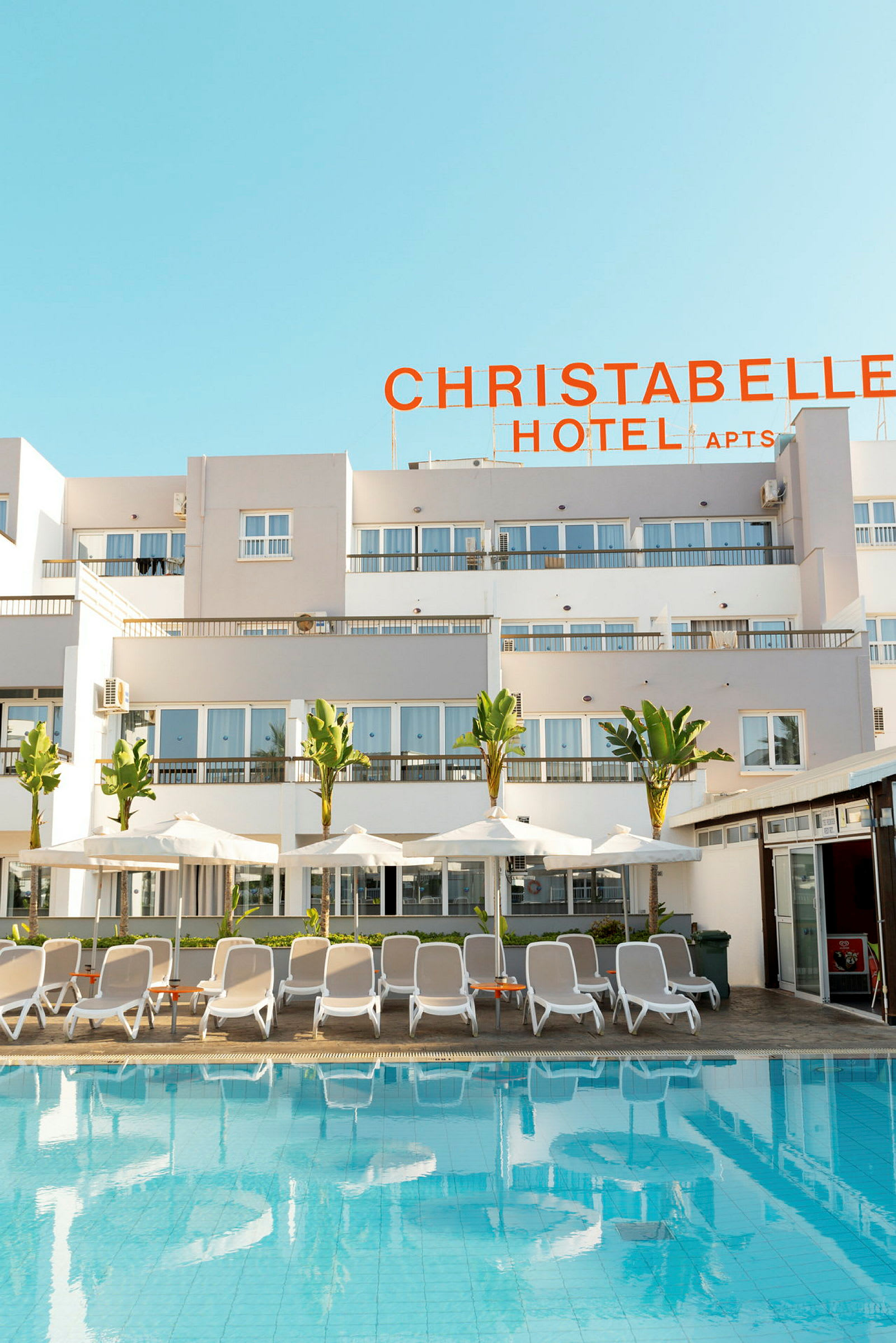 Utendørsbasseng med solsenger og parasoller foran Christabelle Hotel. (AI-generert tekst)