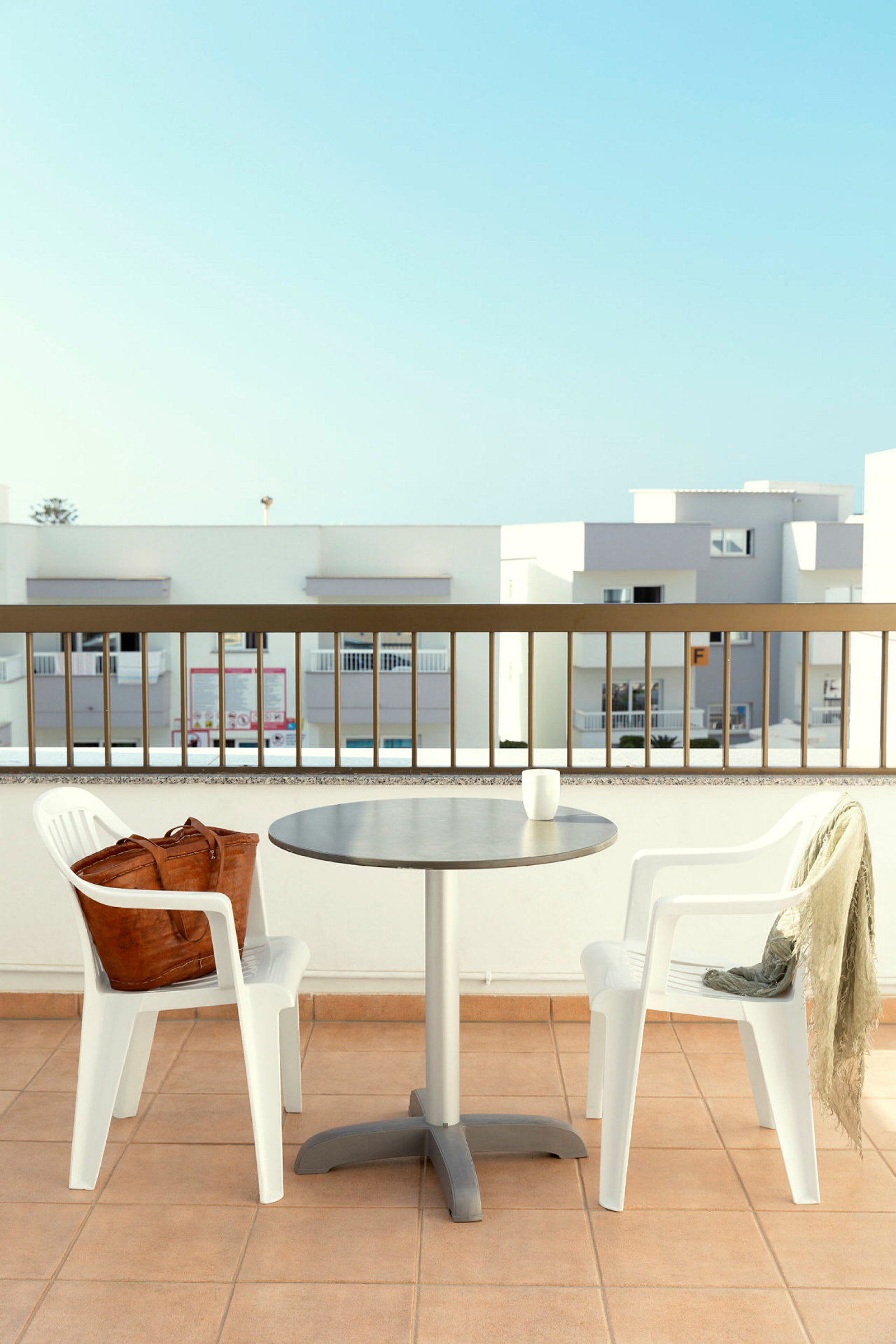 To hvite plaststoler og et bord på en terrasse med rekkverk og moderne byggearkitektur i bakgrunnen. (AI-generert tekst)