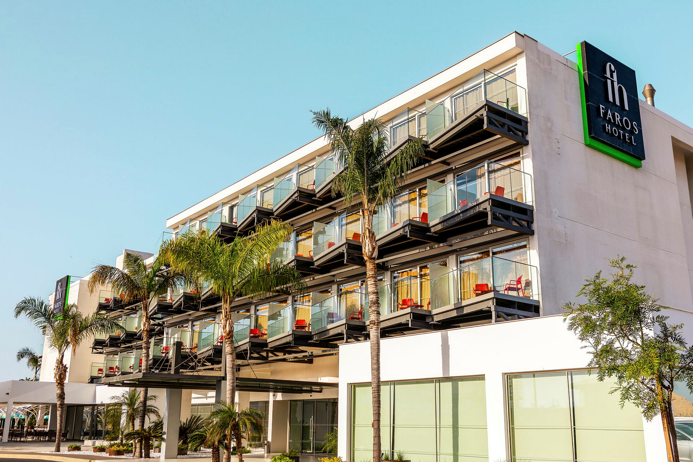 Utendørsbilde av Faros Hotel med flere etasjer, glassbalkonger og palmetrær foran. (AI-generert tekst)