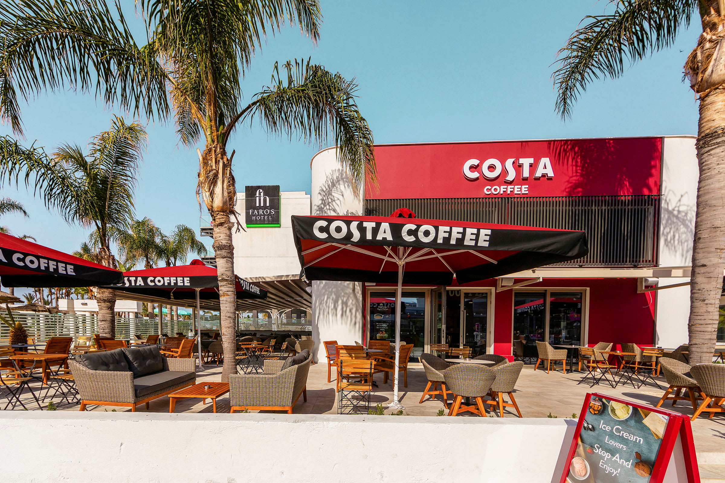 Utendørsservering ved caféen Costa Coffee med palmer og sittemøbler. (AI-generert tekst)