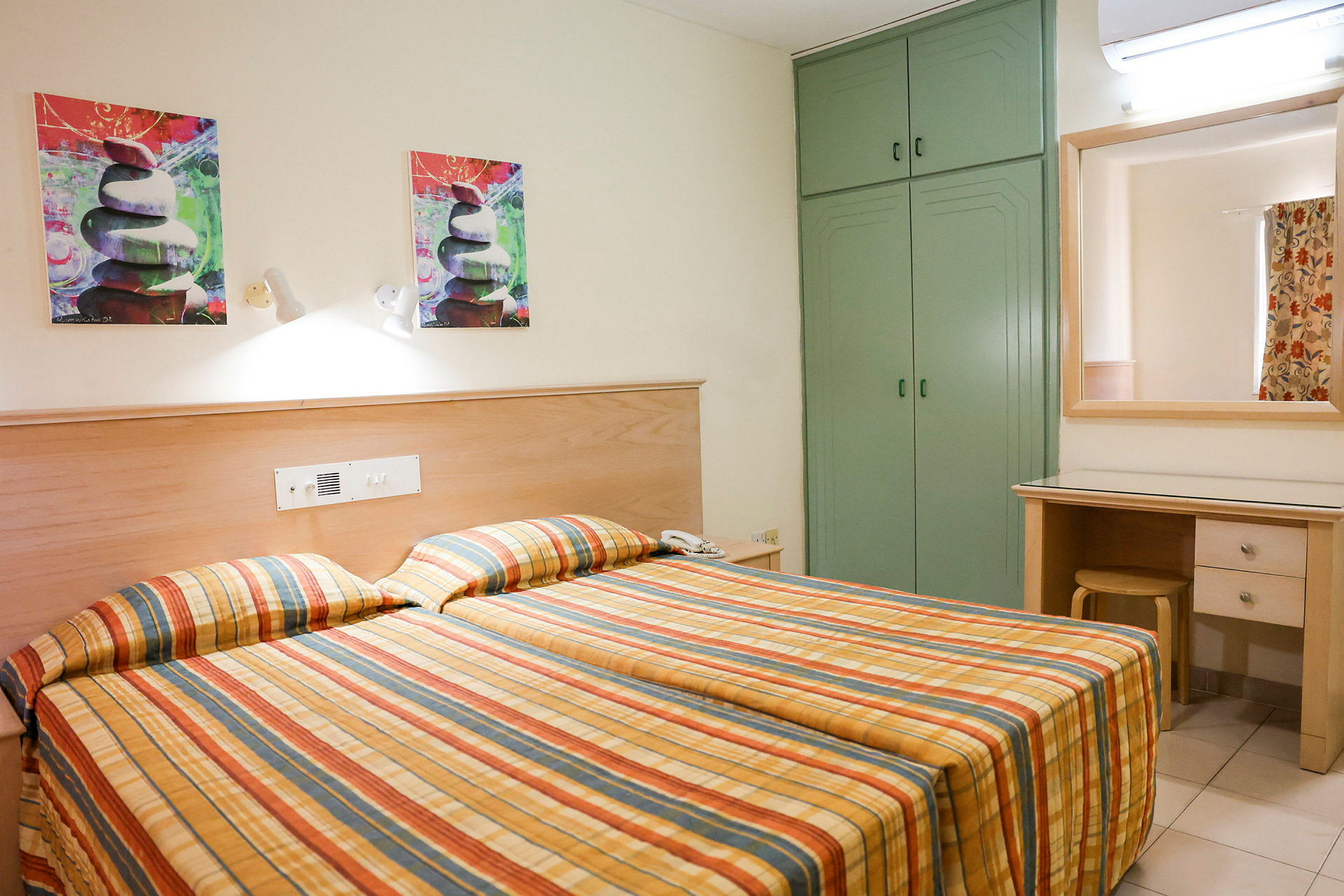 Hotellrom med to enkeltsenger og rutete sengetepper. Det er et grønt skap, et speil over et skrivebord og kunstmalerier på veggen. (AI-generert tekst)