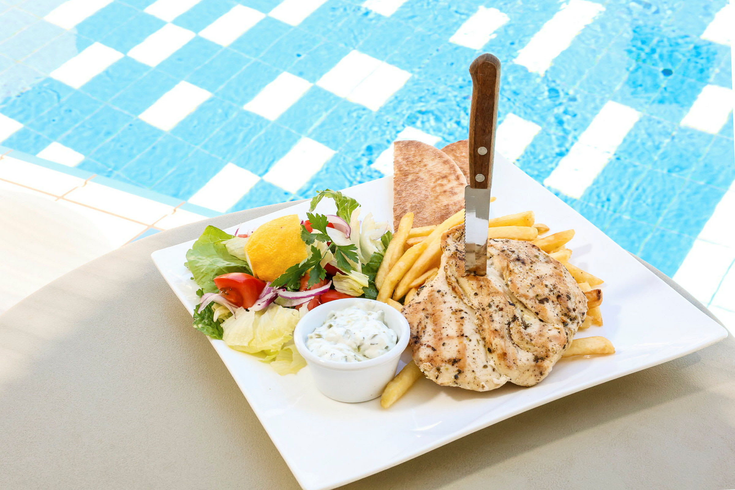 En tallerken med kylling, pommes frites, pitabrød og salat ved siden av et basseng. (AI-generert tekst)