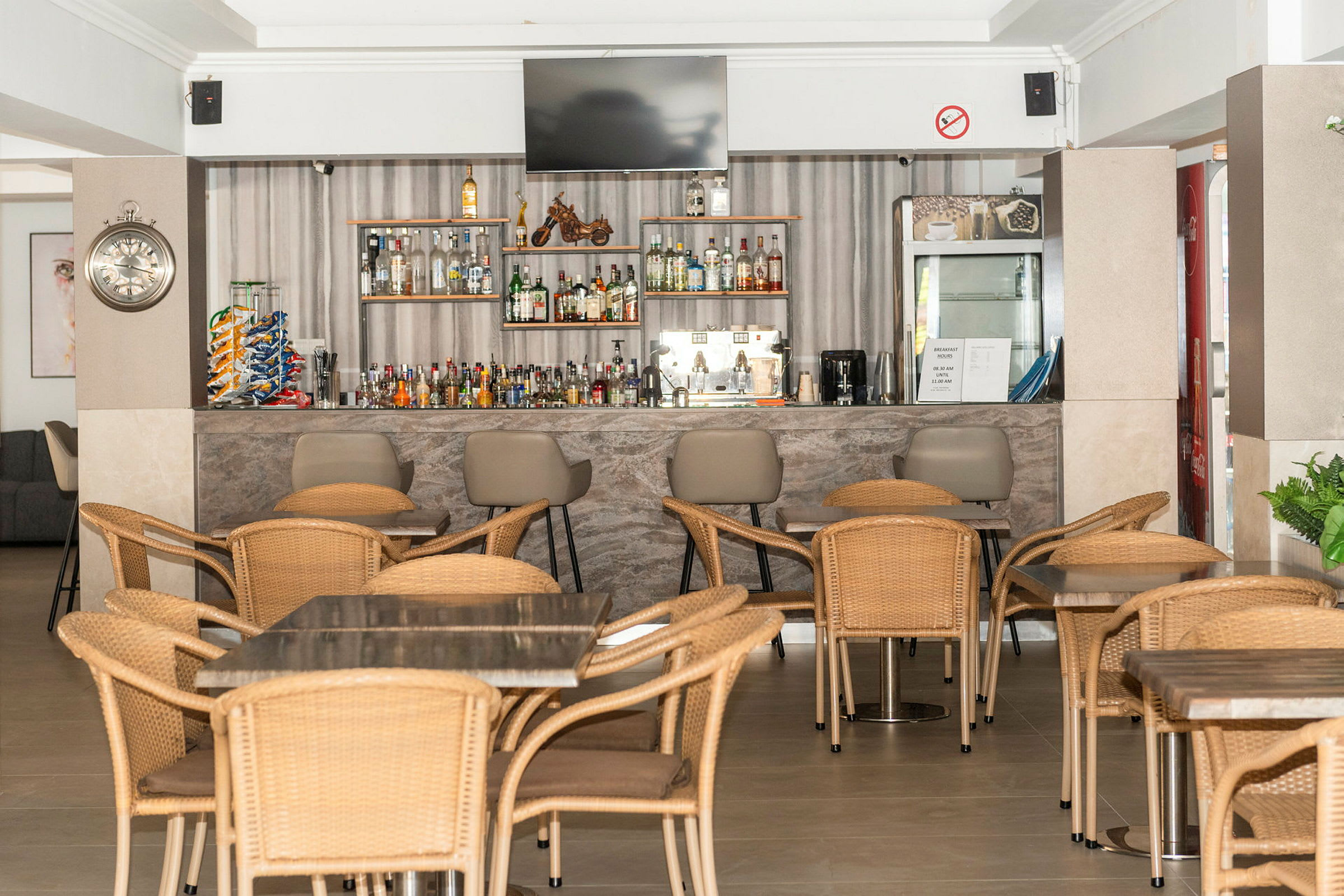 En hotellbar med bord og stoler av rotting og en velutstyrt hylle med drikkevarer. (AI-generert tekst)