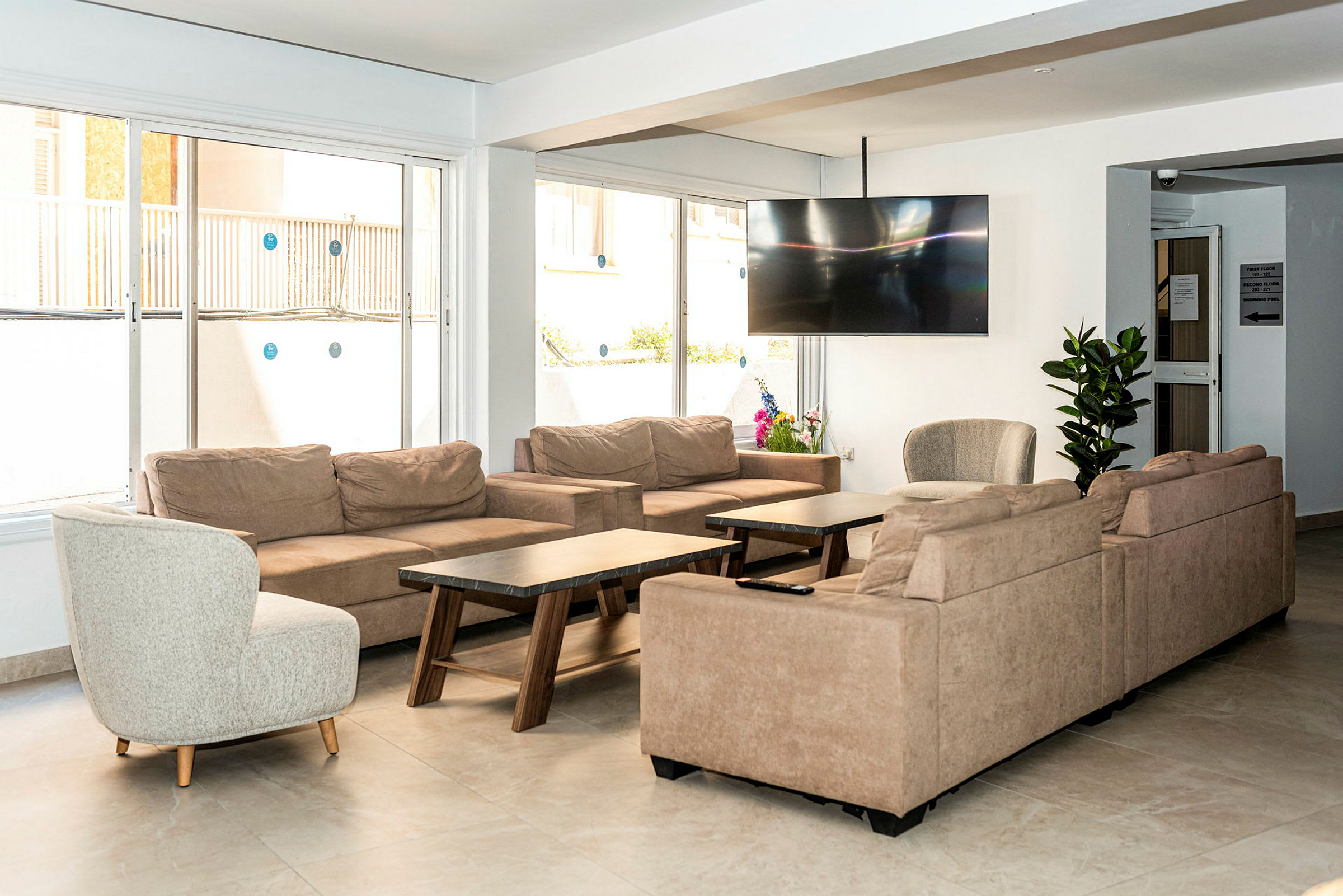 En stue med beige sofaer, en rund lenestol, sofabord og en stor flatskjerm-TV på veggen samt planter til dekorasjon. (AI-generert tekst)