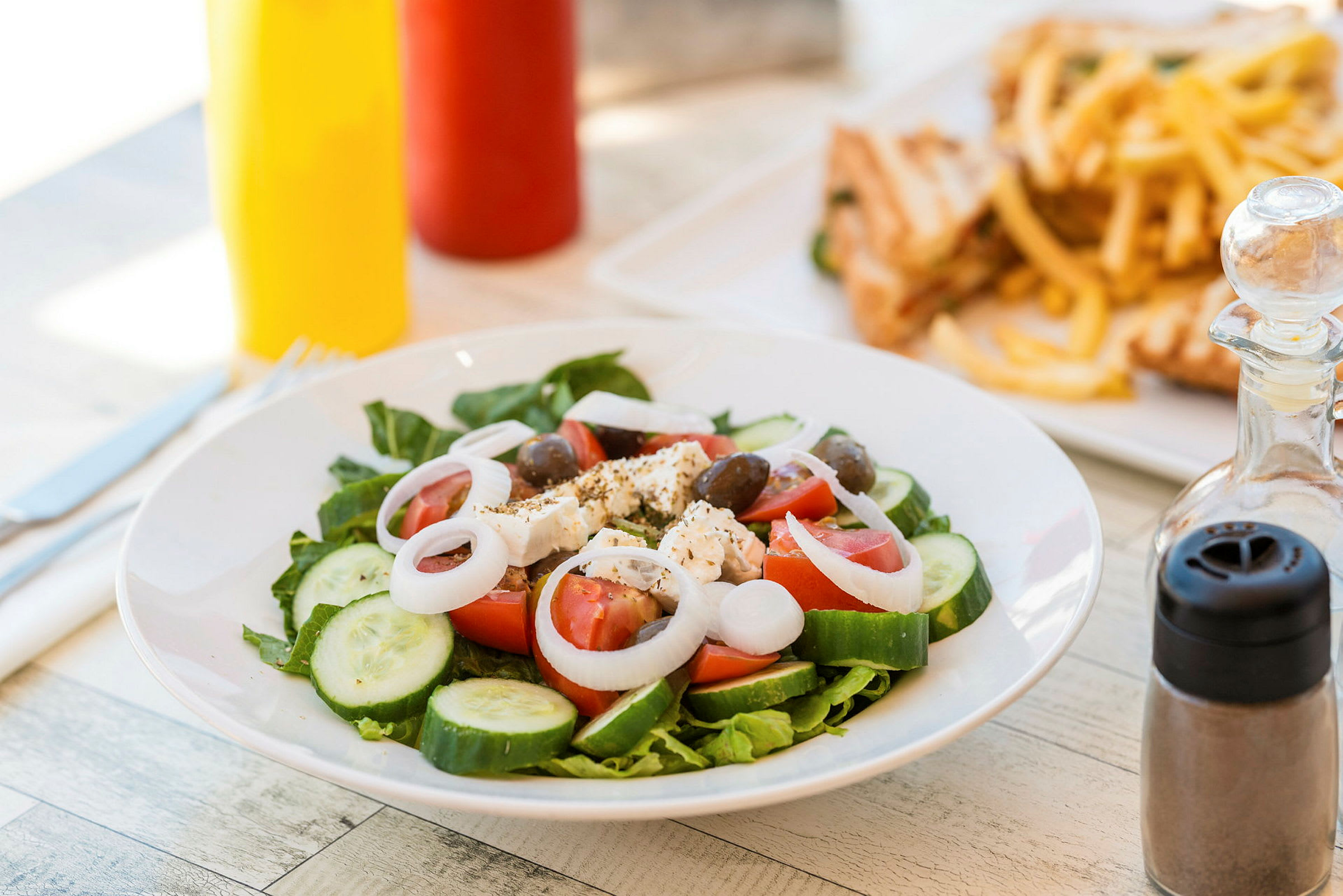 En tallerken med salat som inneholder tomat, agurk, løk, fetaost og oliven. I bakgrunnen sees pommes frites og smørbrød. (AI-generert tekst)