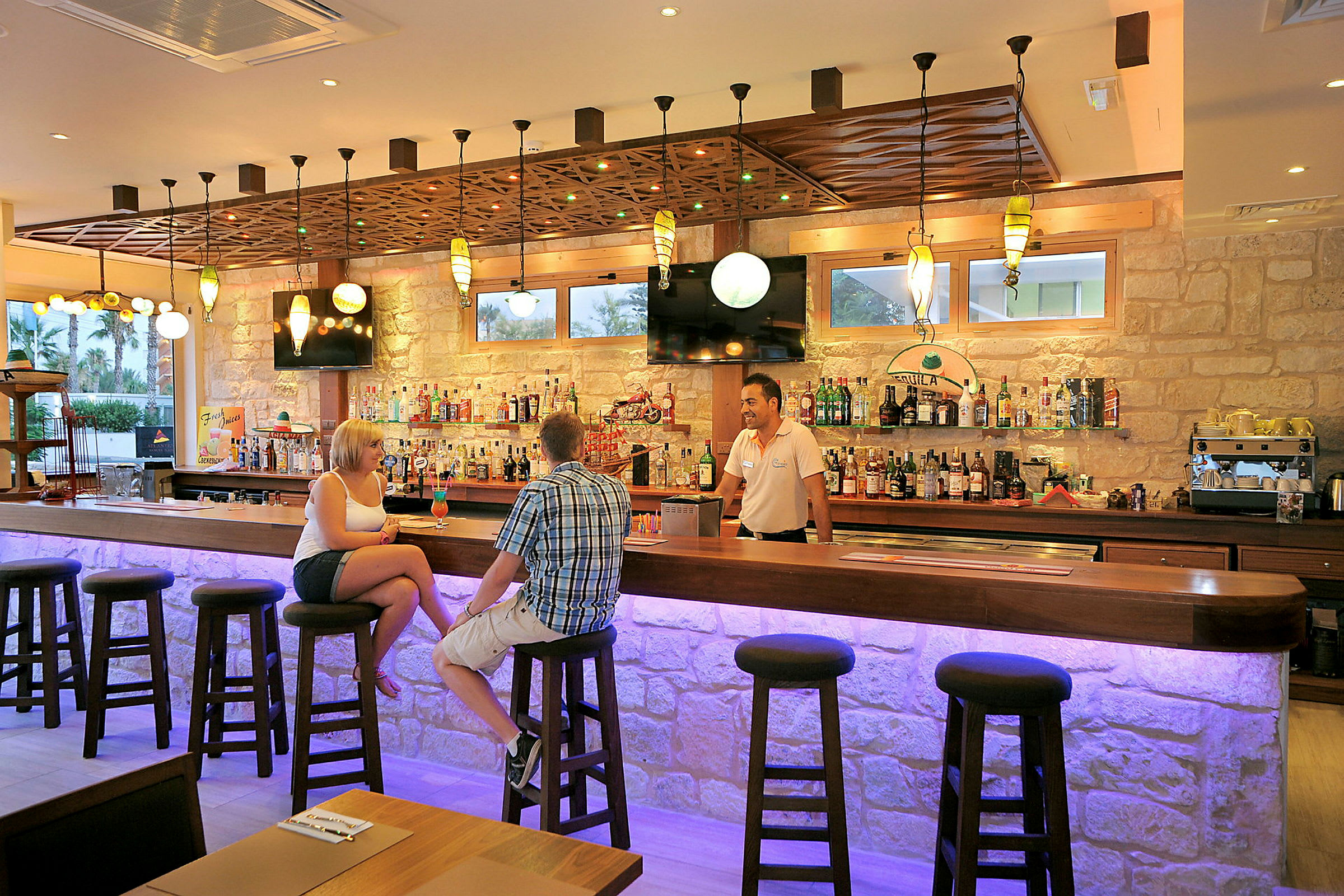 Interiør av en bar med en opplyst bardisk og mennesker som sitter på barkrakker. (AI-generert tekst)