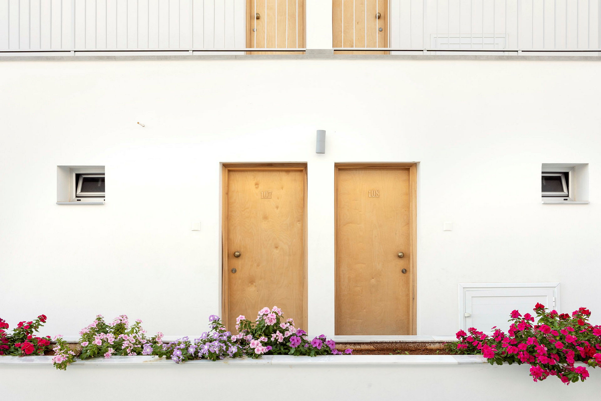 To tredører med nummer 107 og 108 på en hvit vegg, omgitt av flerfargede blomster utenfor. (AI-generert tekst)