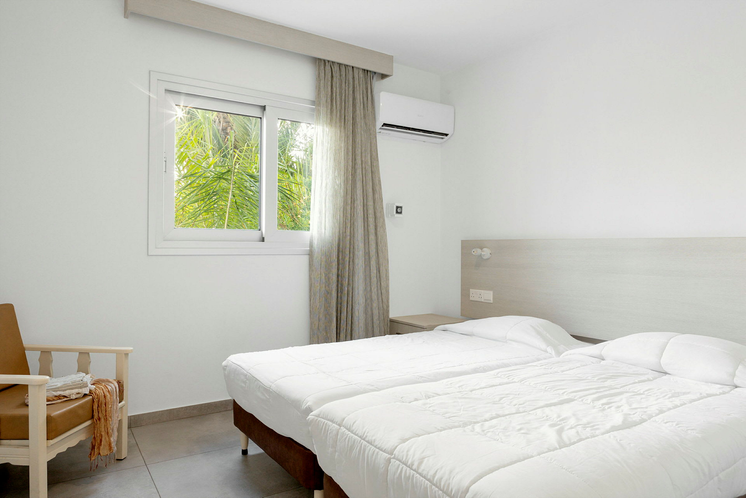 Hotellrom med to enkeltsenger, en lenestol og et vindu med utsikt mot grøntområder. (AI-generert tekst)