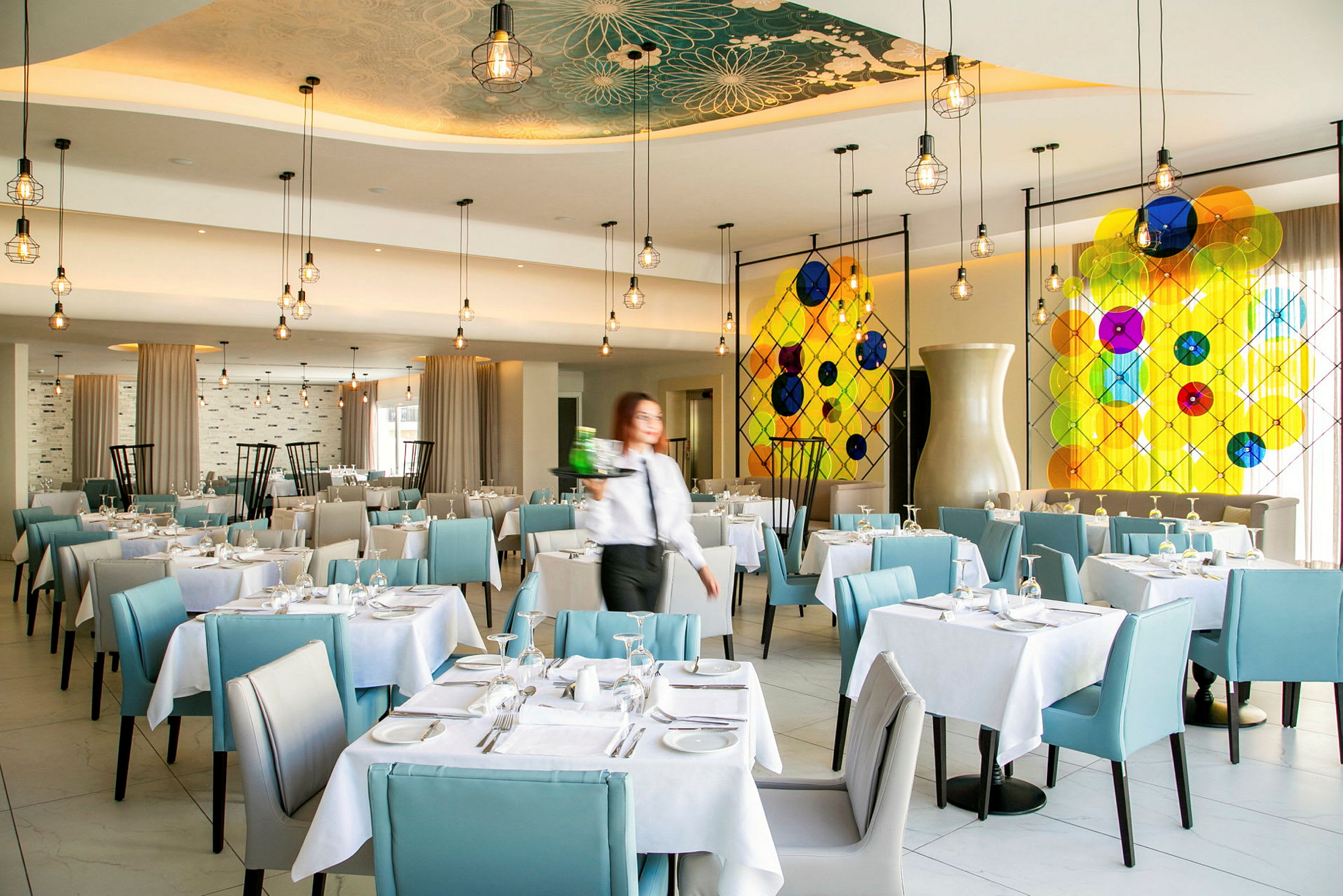 Restaurant med oppdekkede bord og blå stoler samt fargerik glassdekorasjon på veggen. (AI-generert tekst)