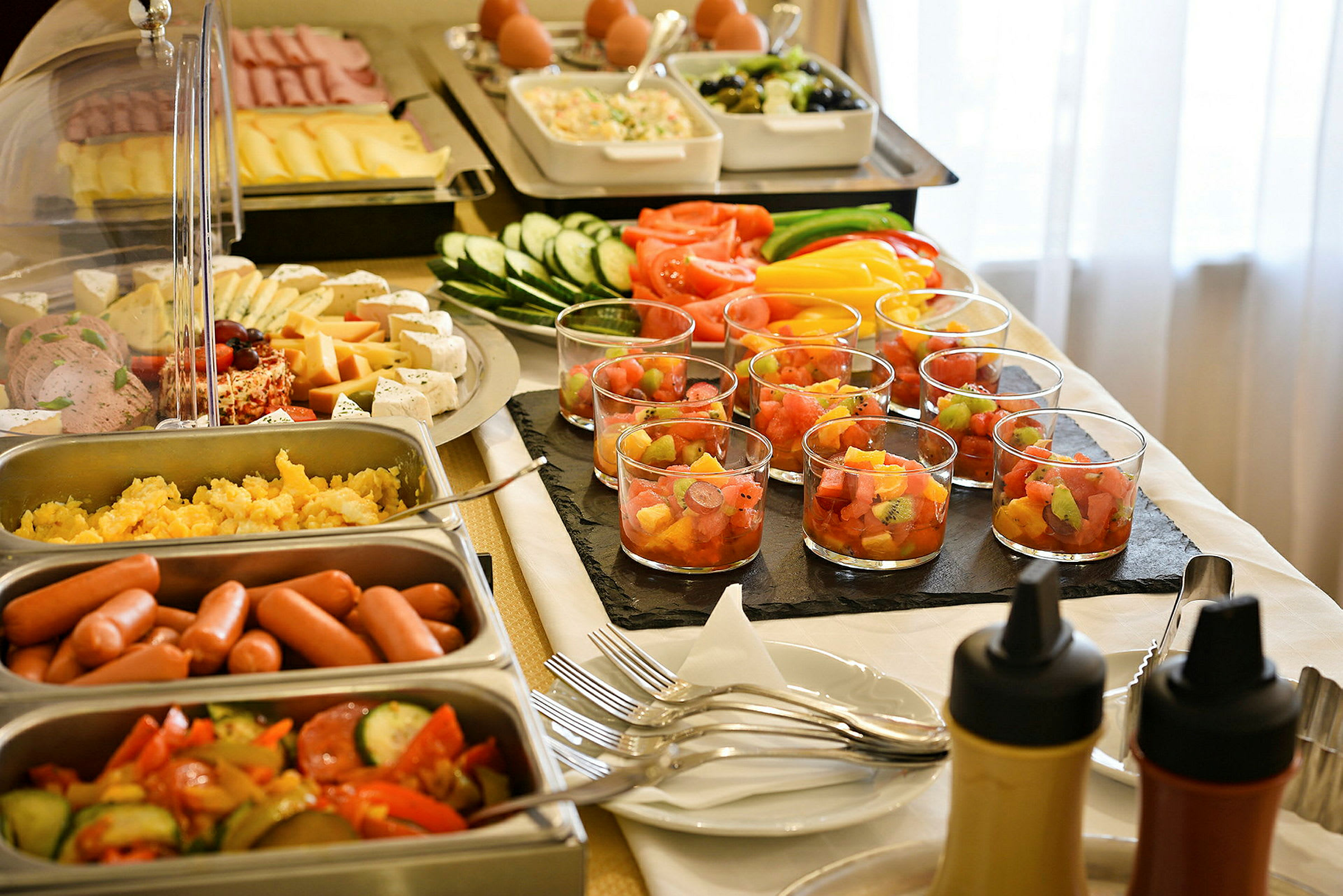 En buffet med diverse mat som pølser, grønnsaker, ost, fruktsalat i glass og sauser. (AI-generert tekst)