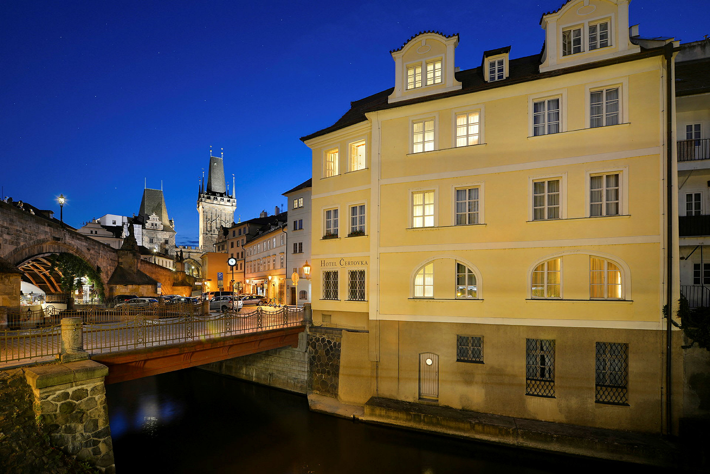 En liten bro over en kanal ved siden av en gul bygning under en blå nattehimmel. I bakgrunnen sees en historisk steinbro og en kirkespirelignende bygning. (AI-generert tekst)