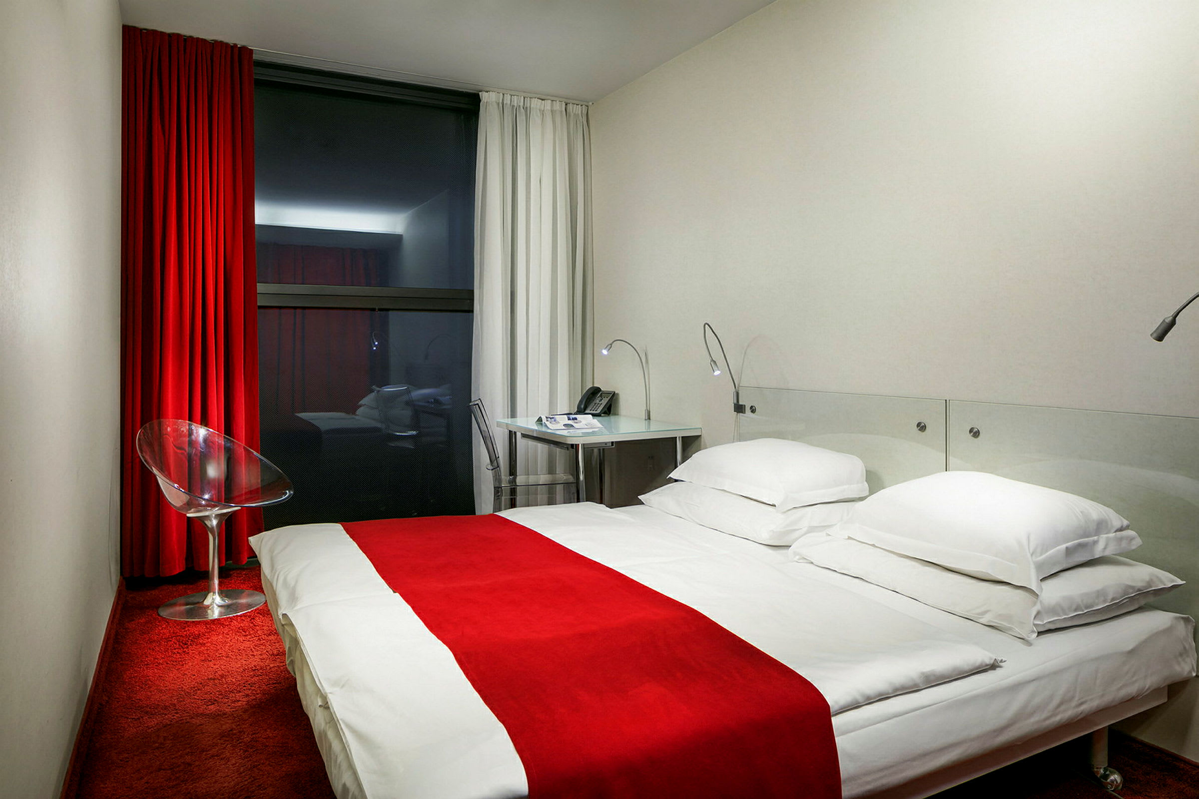 Hotellrom med rødt teppe, hvit seng med rødt sengeteppe, glassstol og skrivebord. (AI-generert tekst)