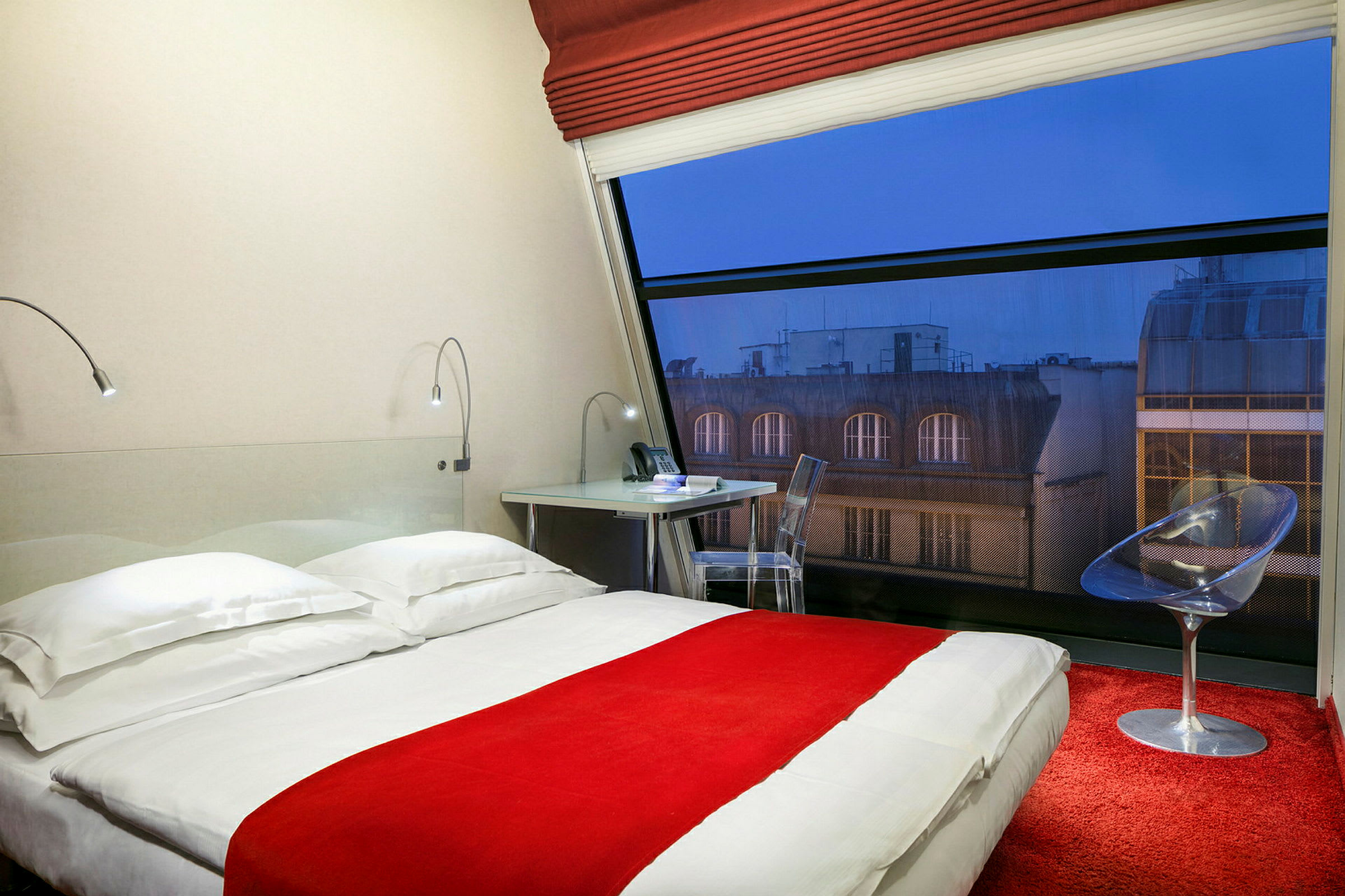 Hotellrom med en seng, skrivebord og stol samt et stort vindu med utsikt. (AI-generert tekst)