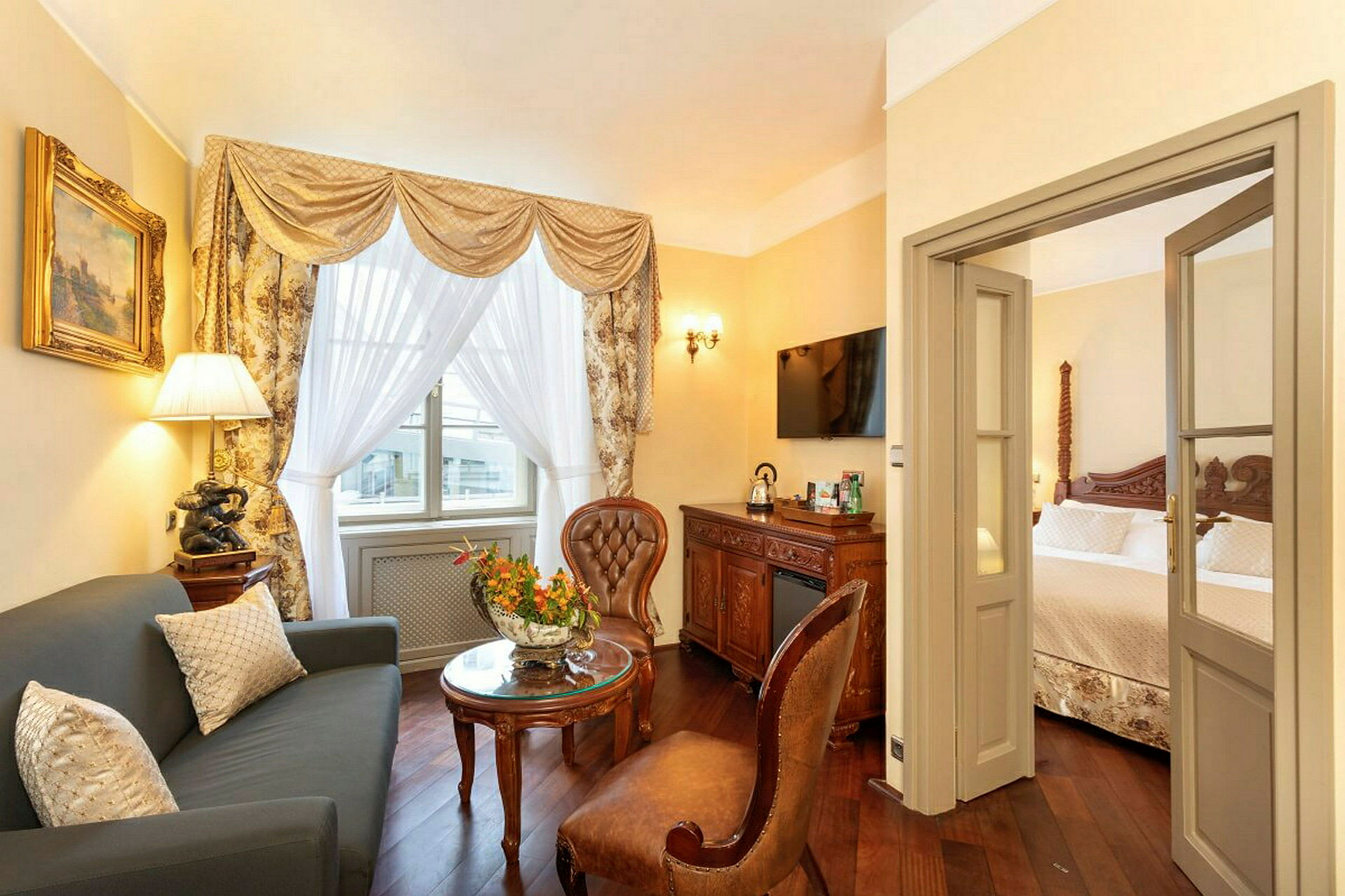 Et elegant innredet hotellrom med en stue med sofa og stoler samt et soverom i bakgrunnen. Det er dekorative gardiner ved vinduet og en døråpning mellom rommene. (AI-generert tekst)