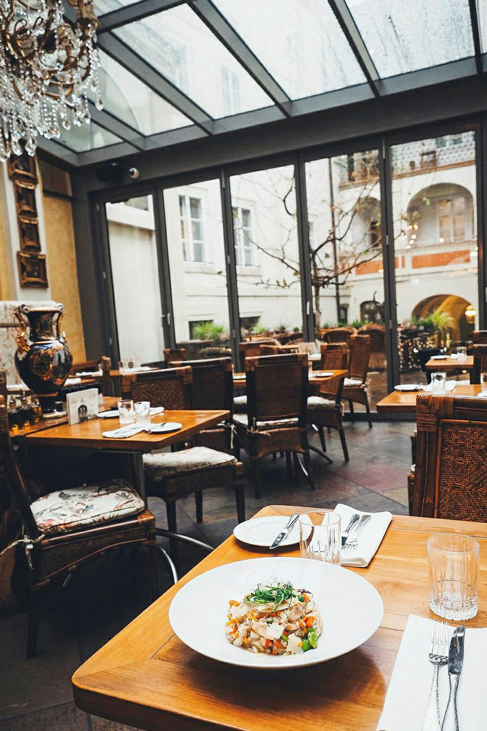 Et restaurantbord er dekket med en tallerken mat og bestikk, plassert i et stilfullt innredet rom med store glassvinduer som gir utsikt til en gårdsplass. (AI-generert tekst)