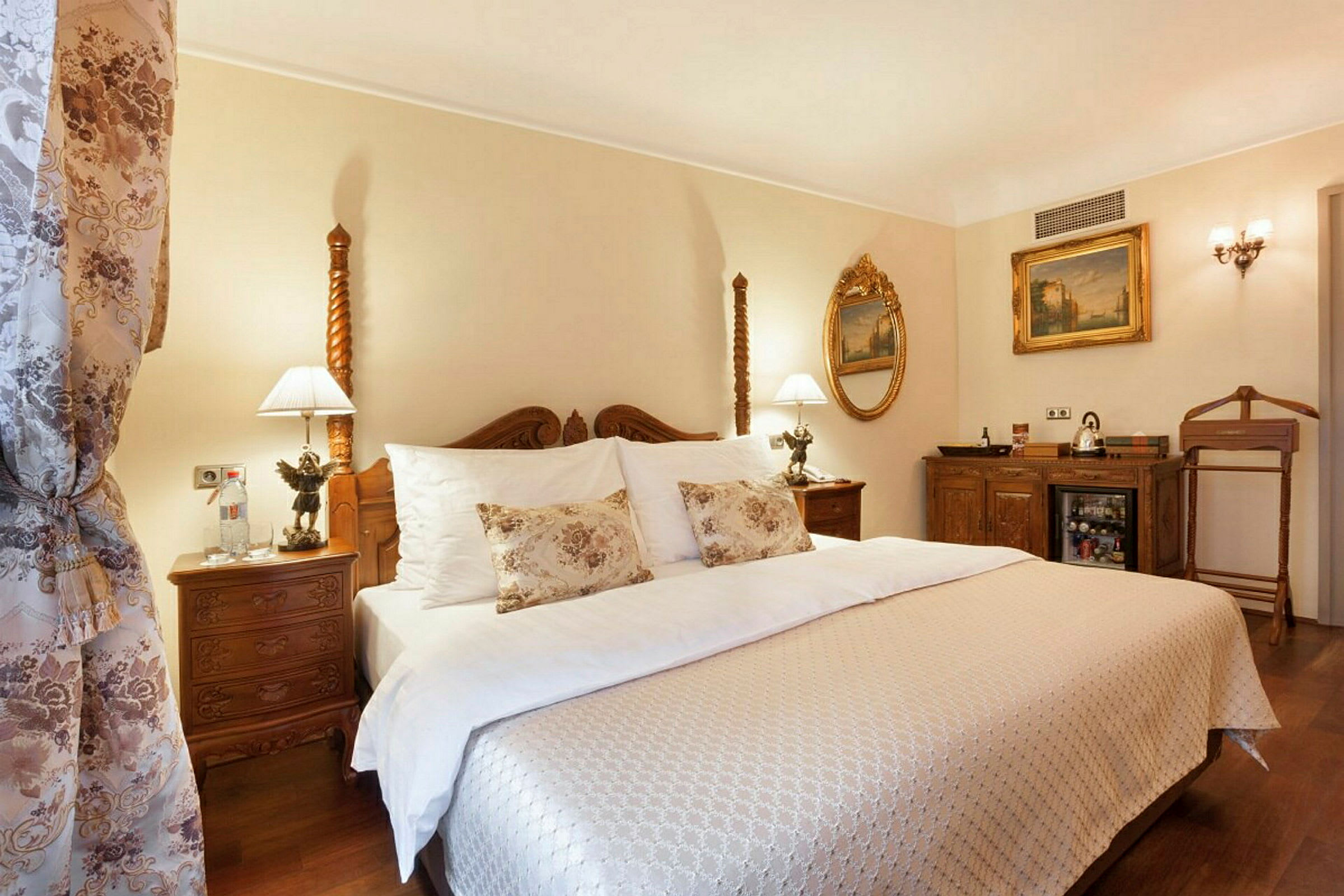 Et elegant hotellrom med en dobbeltseng kledd i hvitt og mønstrede tekstiler, dekorasjoner i klassisk stil og lamper på nattbordene. (AI-generert tekst)
