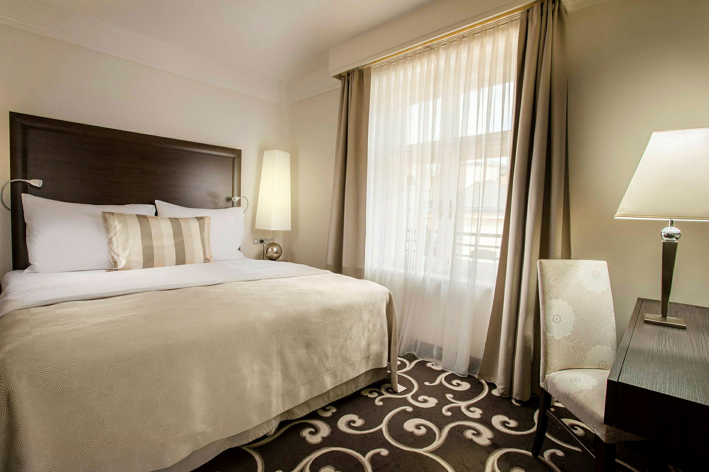 Et hotellrom med en stor seng med beige og hvitt sengetøy, et teppe med mønster, et skrivebord med en bordlampe og en stol ved siden av. Vinduet har beige gardiner og hvit gjennomskinnelig tyll. (AI-generert tekst)