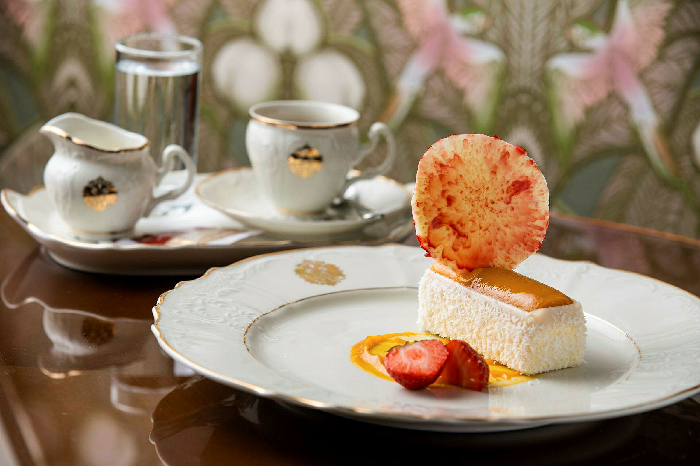 En dessert på en tallerken med dekorasjoner. Bak er det en kaffekopp og en fløtemugge. (AI-generert tekst)