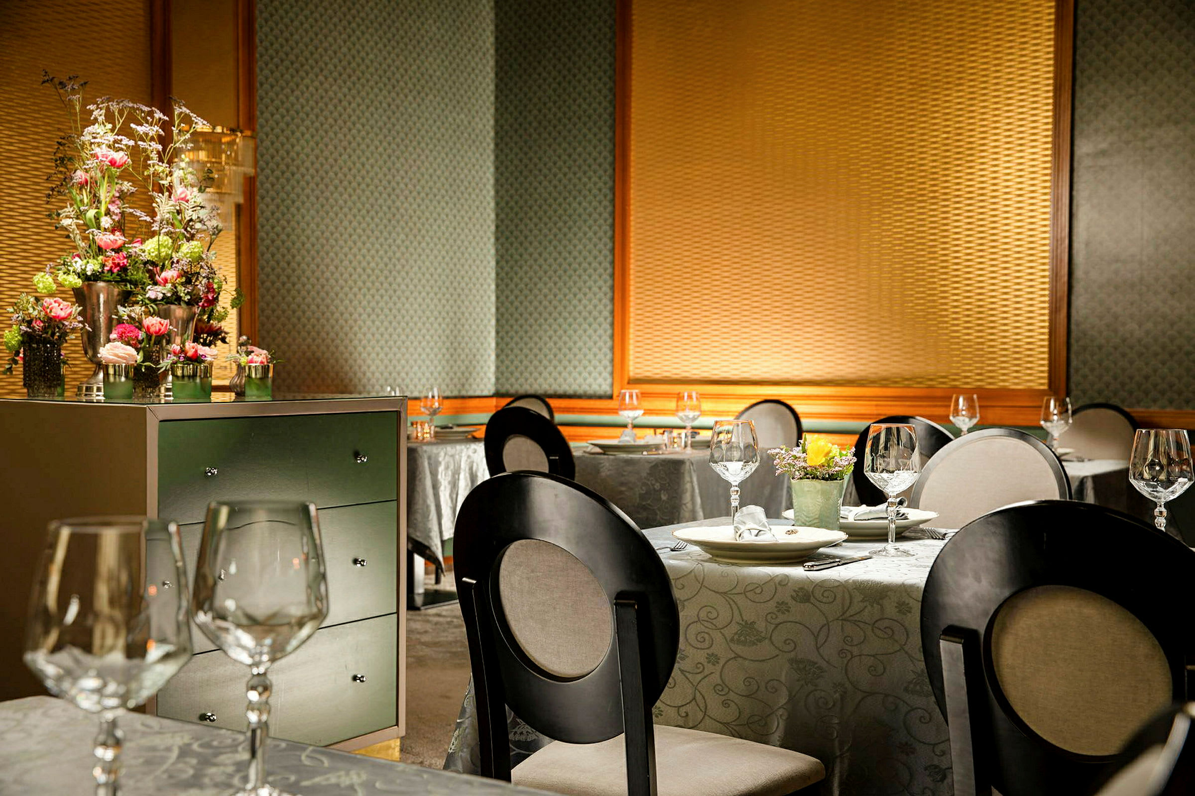 En elegant restaurantinteriør med dekte bord, glass og stoler. Et skap med blomsterarrangement er synlig i forgrunnen. (AI-generert tekst)