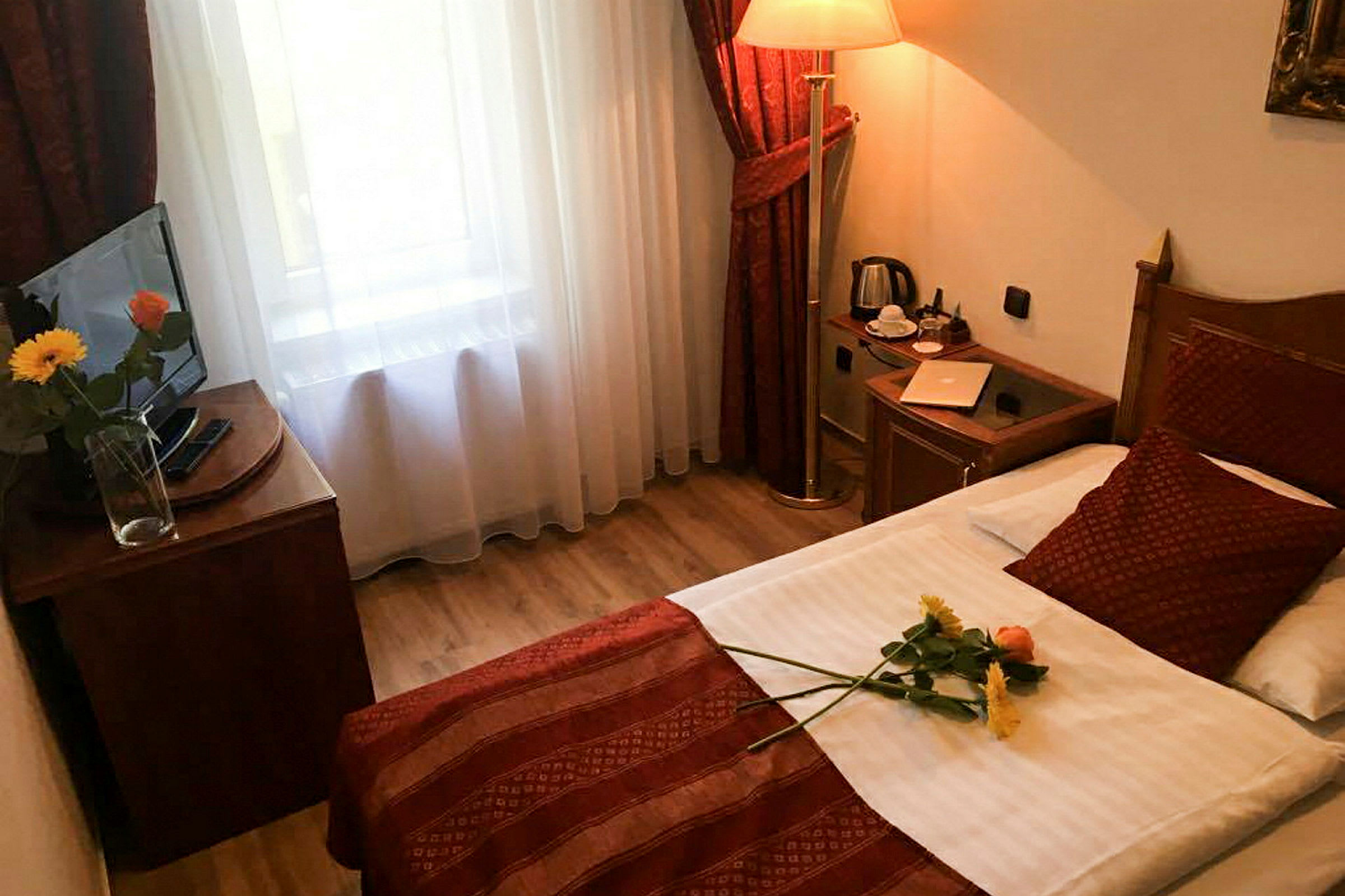 Et hotellrom med en enkeltseng, et nattbord med kopp og vannkoker, samt en TV på en benk ved siden av et vindu. En vase med blomster står på TV-benken, og en blomst ligger på sengen. (AI-generert tekst)