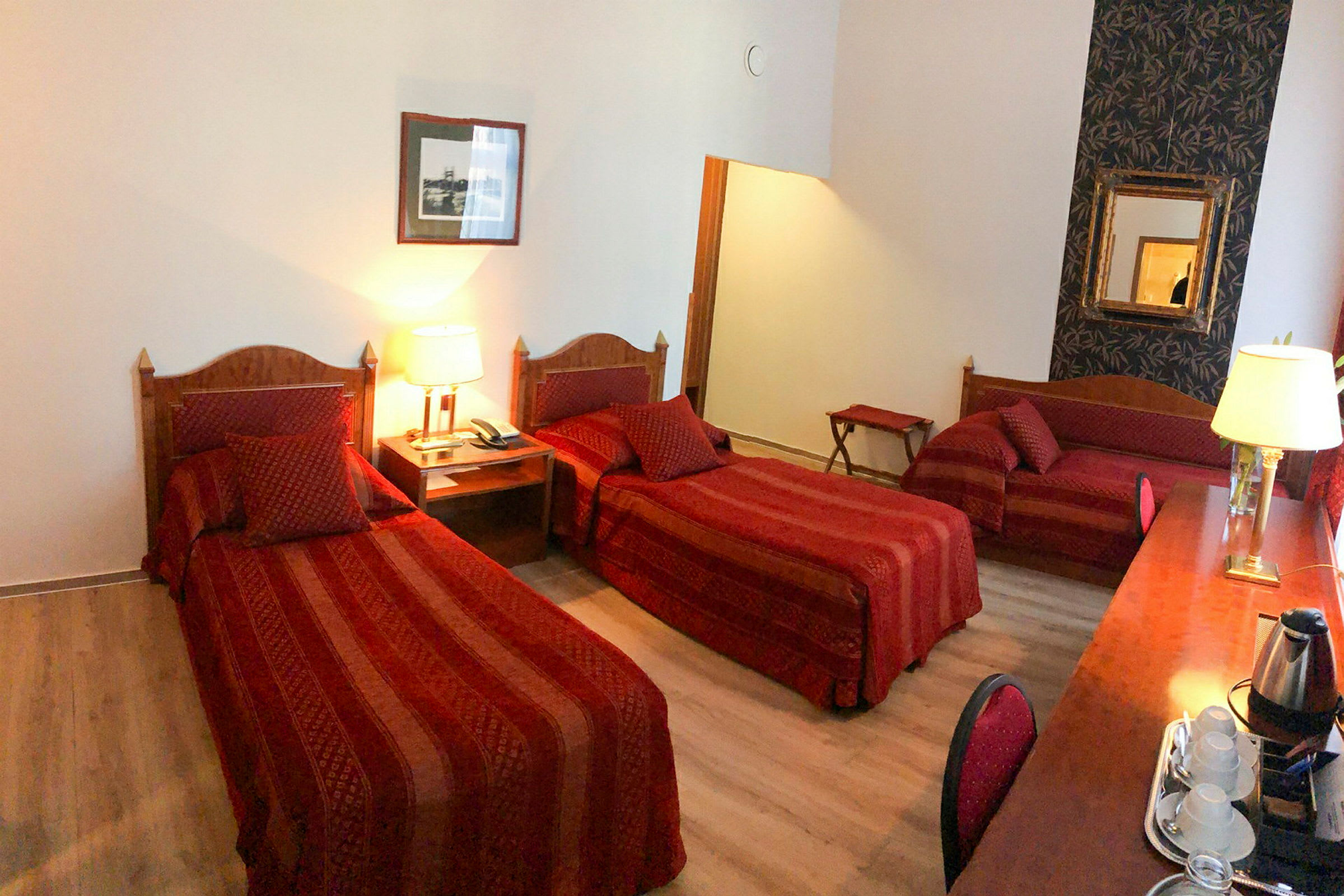 Hotellrom med tre senger i et romslig innredet område, dekorert med røde tekstiler og tredetaljer. (AI-generert tekst)