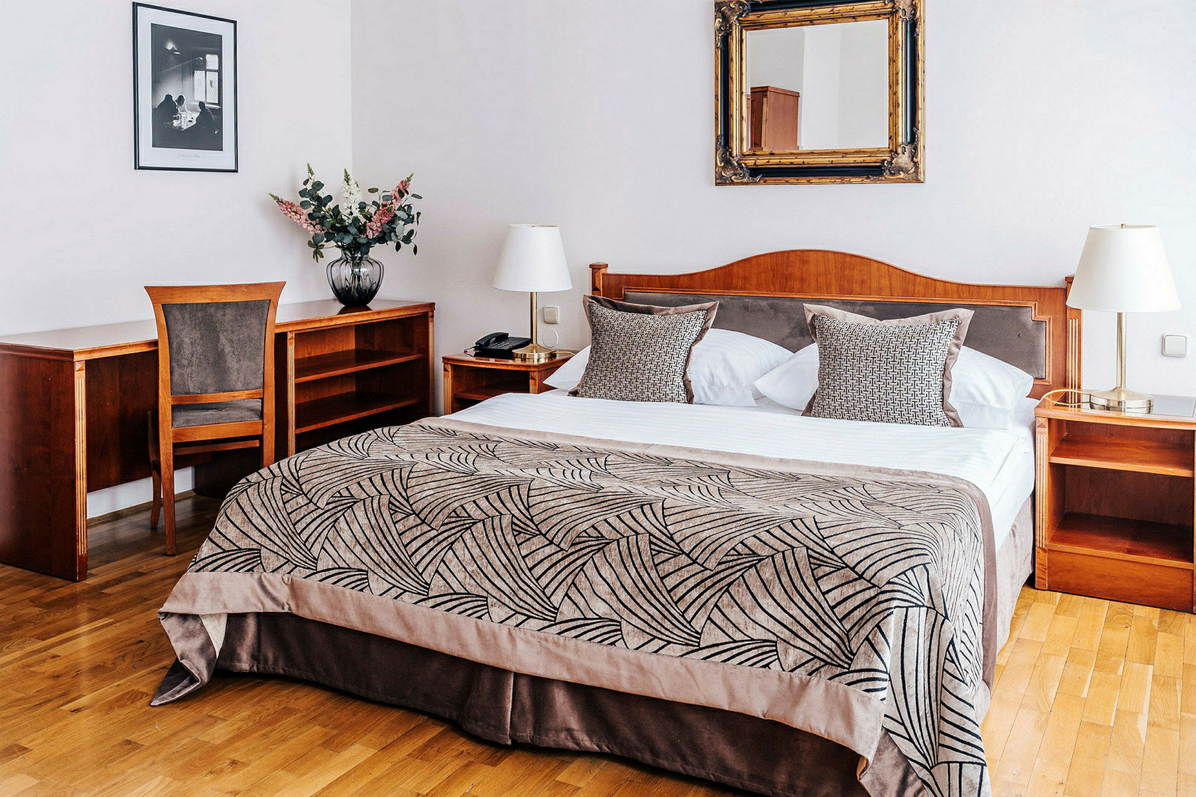 Et hotellrom med en dobbeltseng, nattbord og skrivebord av tre. På sengen ligger et mønstret sengeteppe og puter. (AI-generert tekst)