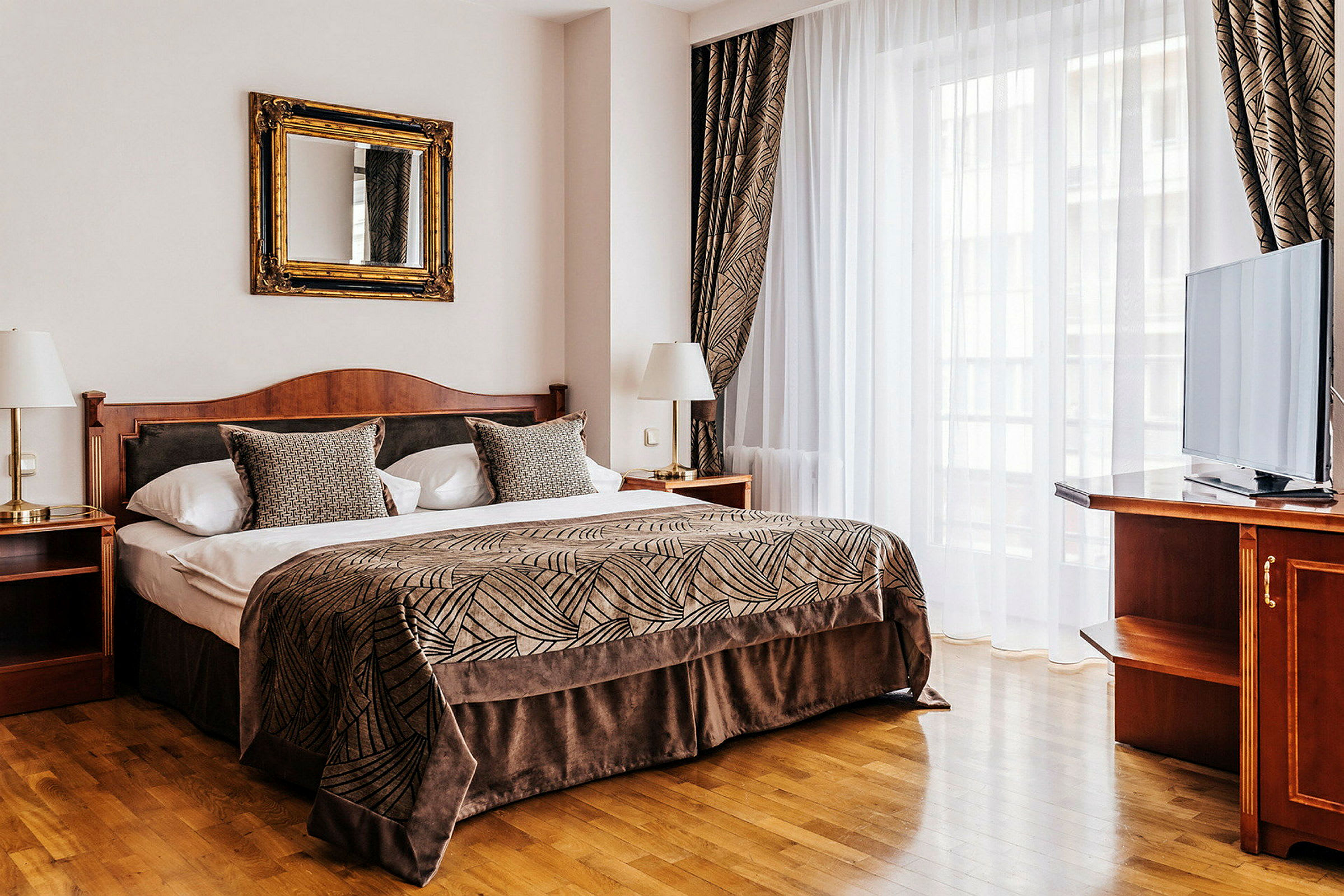 Et elegant hotellrom med en stor seng, dekket med et mønstret sengeteppe og pynteputer. Rommet har et speil over sengen, to nattbord med lamper og en TV på en møbel ved siden av et stort vindu. (AI-generert tekst)
