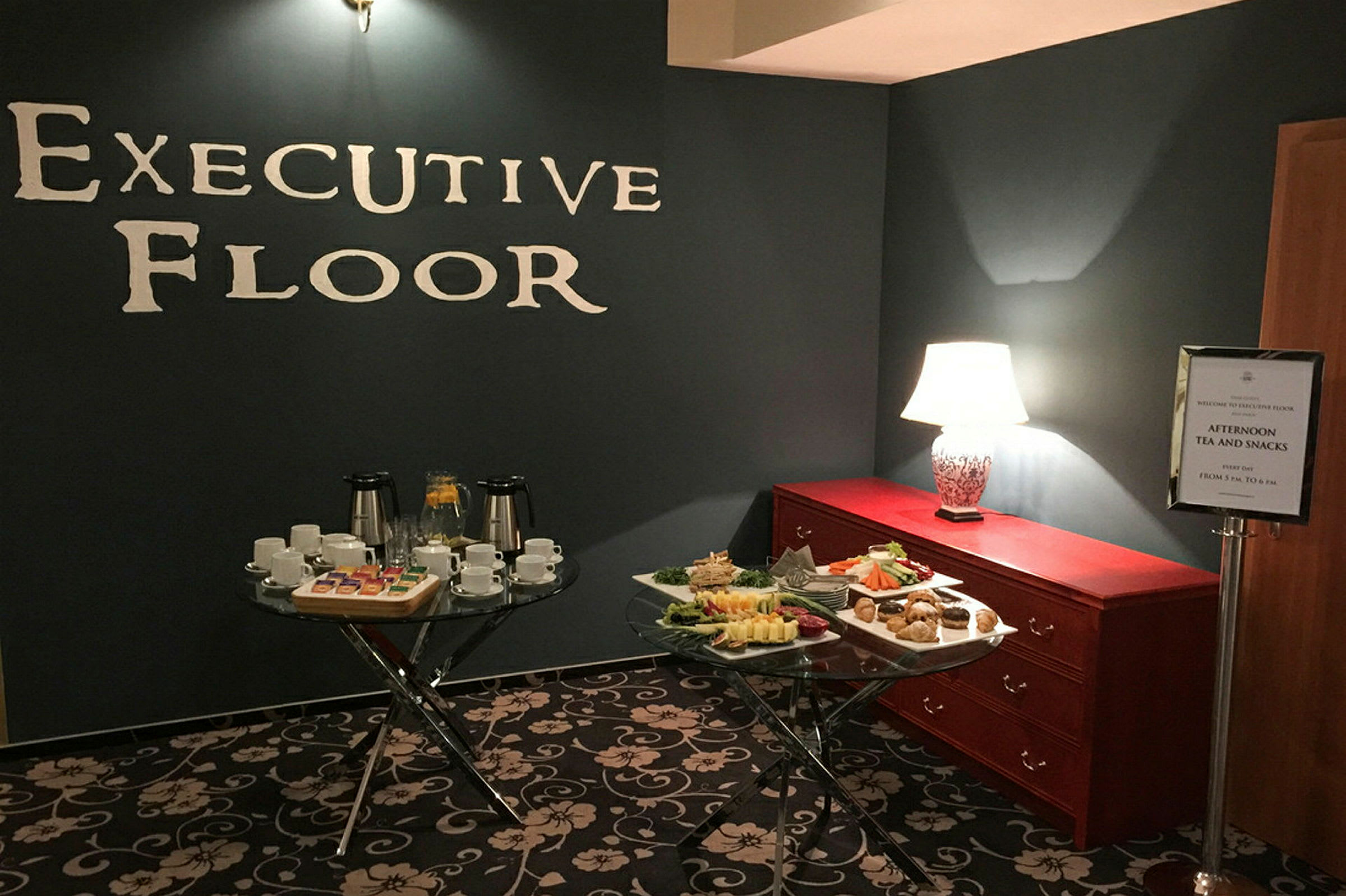 Et rom med teksten "Executive Floor" på veggen, moderne belysning, to bord med kopper, tallerkener og snacks samt en kommode med bordlampe. (AI-generert tekst)