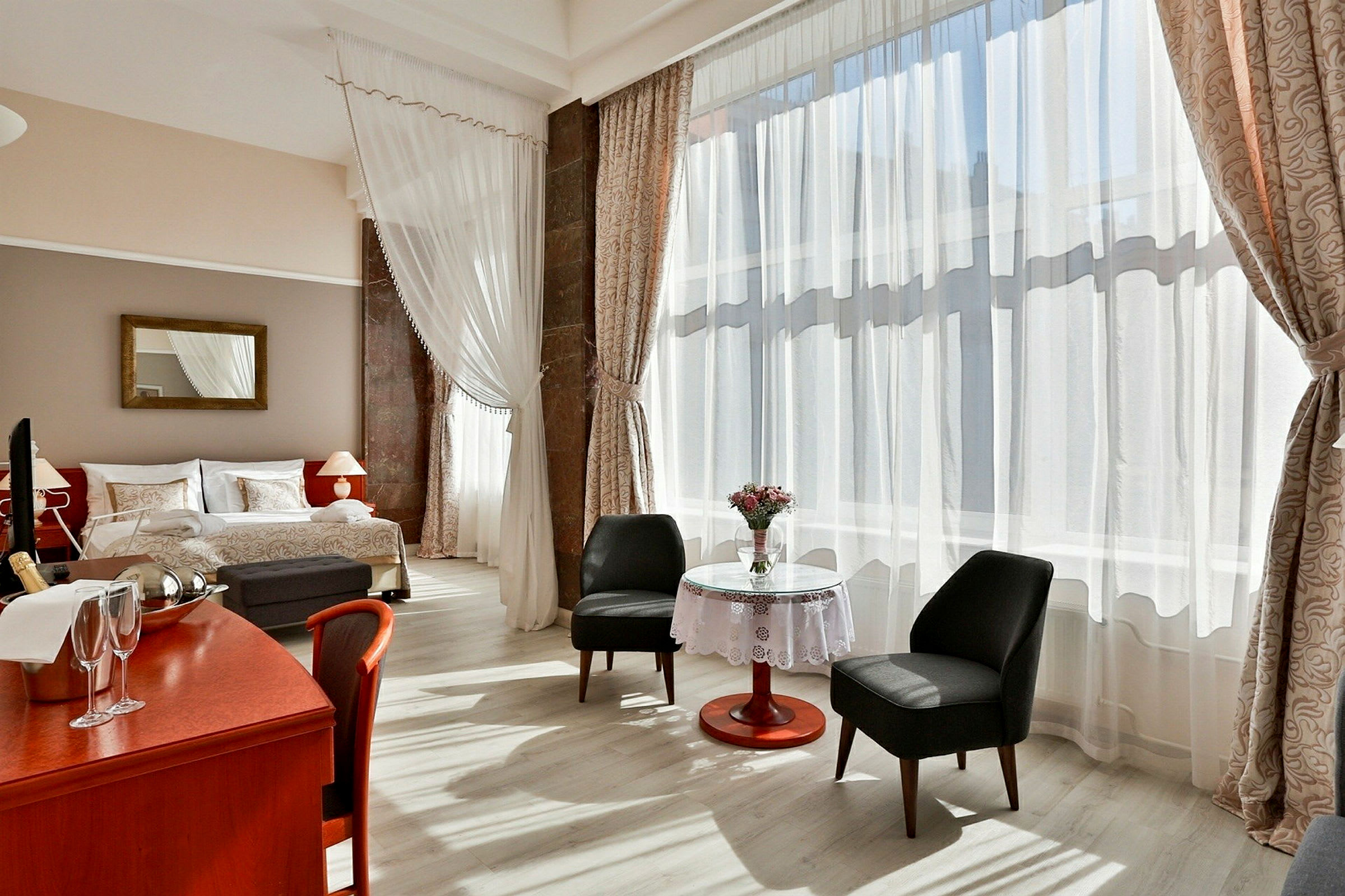 Et hotellrom med store vindusflater og mye dagslys. Rommet har en dobbeltseng, en lesekrok med to stoler og et bord med vase og blomster, samt et skrivebord med champagneflaske og glass. (AI-generert tekst)