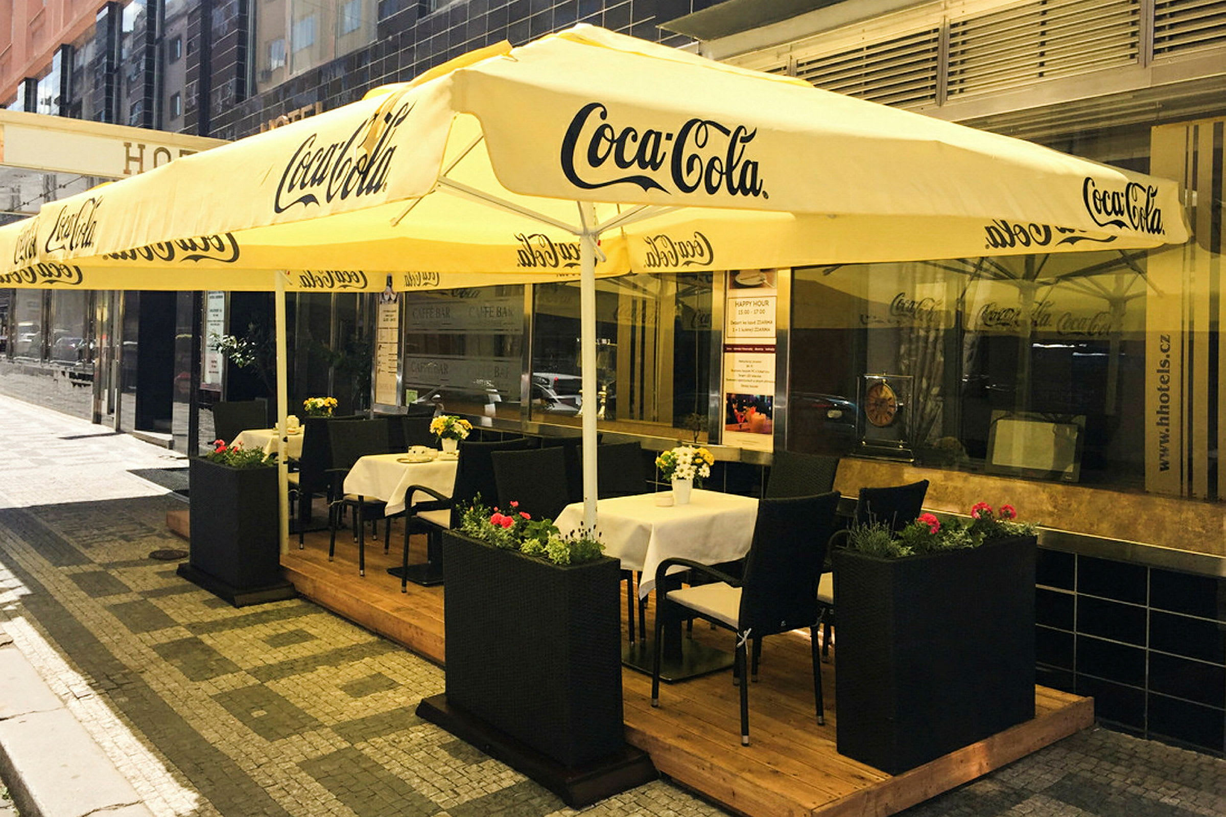 En utekafé med bord og stoler under store gule parasoller merket med Coca-Cola-logoen. (AI-generert tekst)
