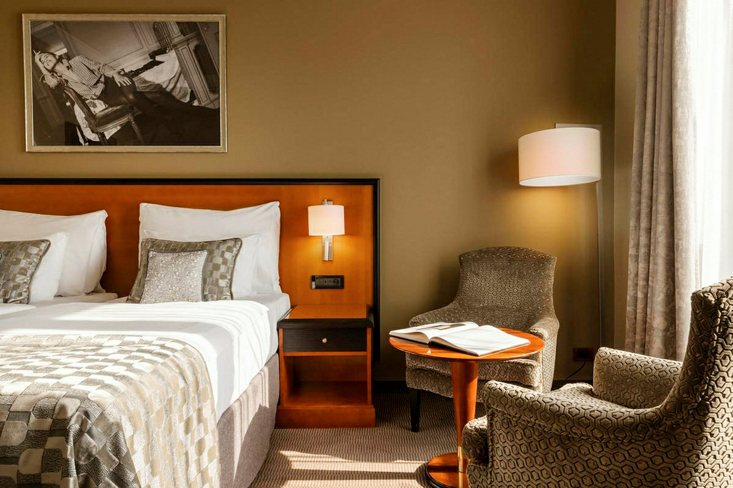 Et hotellrom med en seng, nattbord, lenestoler og et lite bord med en bok på. (AI-generert tekst)