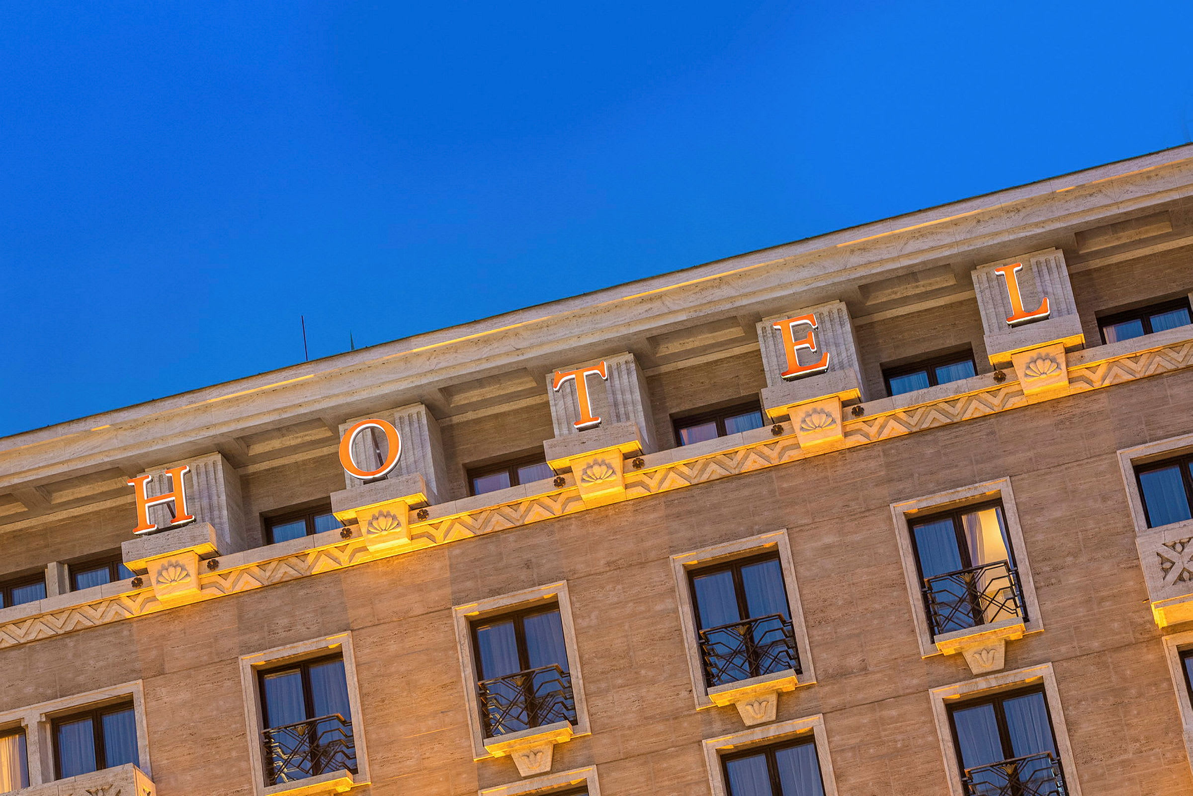 Fasaden på et hotell med opplyste bokstaver som staver ordet 'HOTEL' mot en klarblå kveldshimmel. (AI-generert tekst)