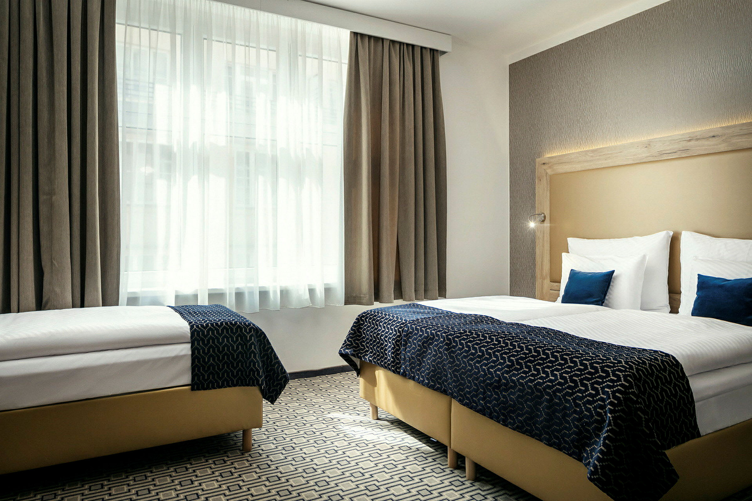 Et hotellrom med to enkeltsenger, hvite laken, blå dekorasjonsputer og et stort vindu med hvite gardiner. (AI-generert tekst)