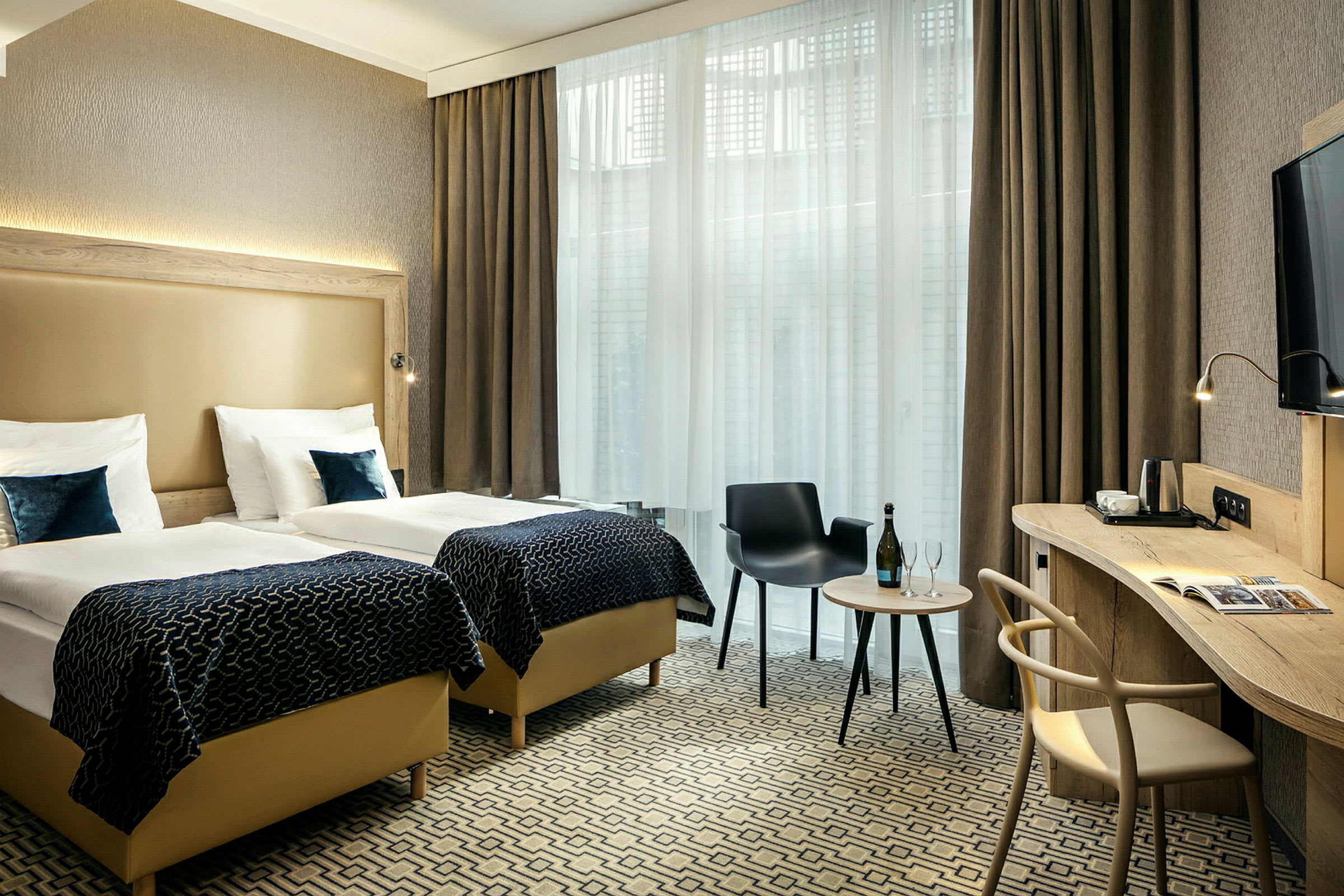 Hotellrom med to enkeltsenger, skrivebord, stol og et lite bord med flaske og glass foran store vinduer med gardiner. (AI-generert tekst)