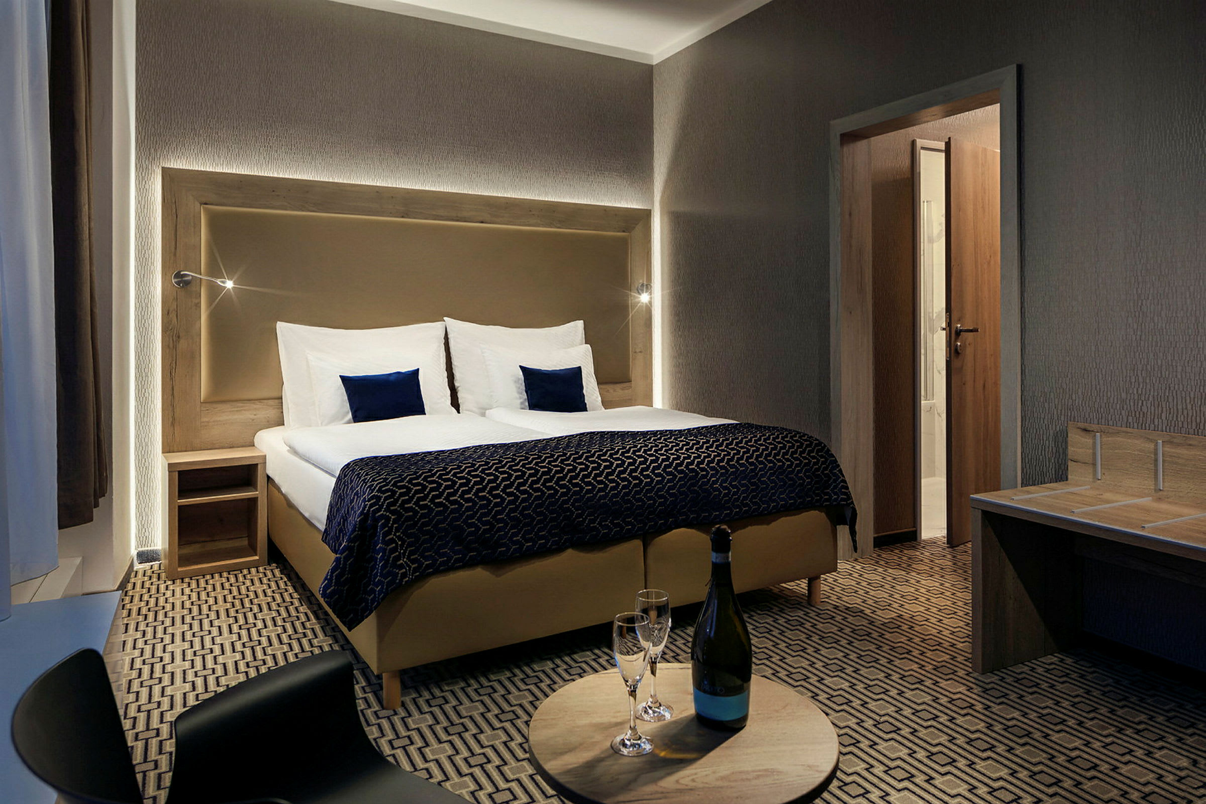 Et hotellrom med dobbeltseng, nattbord og et rundt bord med en flaske og to glass. (AI-generert tekst)