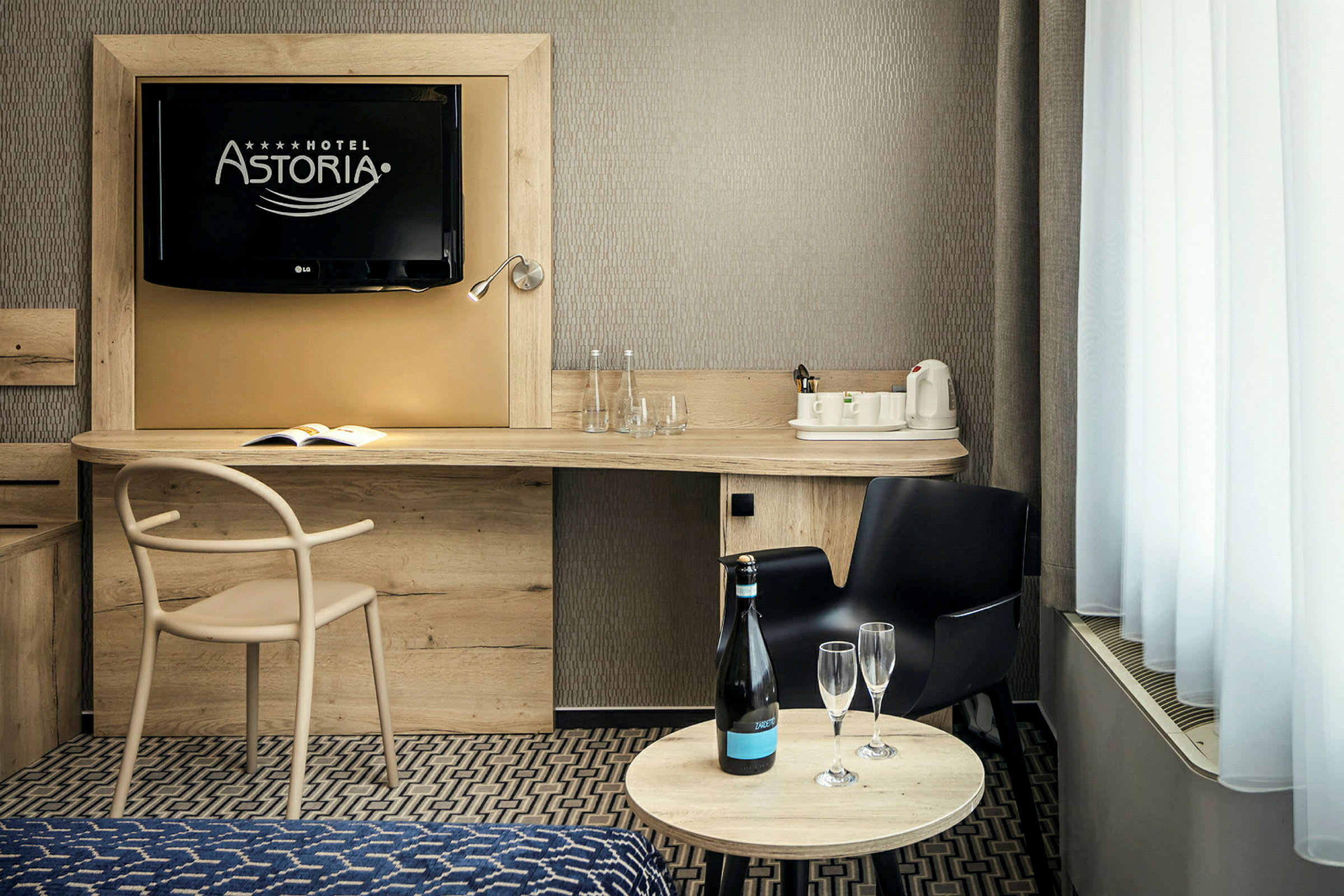 Et hotellrom med en TV på veggen, et skrivebord med en stol, en vannkoker og glass, samt et rundt bord med en flaske og to glass. (AI-generert tekst)
