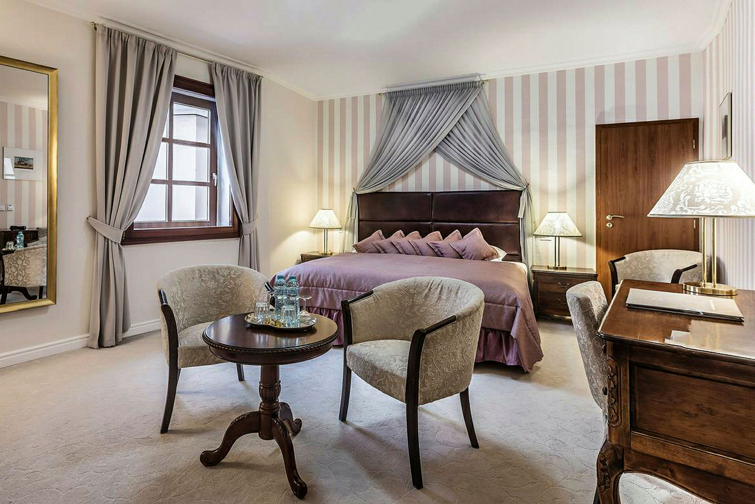 Et elegant hotellrom med en dobbelseng dekket med lilla sengetøy, to lenestoler plassert rundt et lite rundt bord og et skrivebord med lampe. (AI-generert tekst)