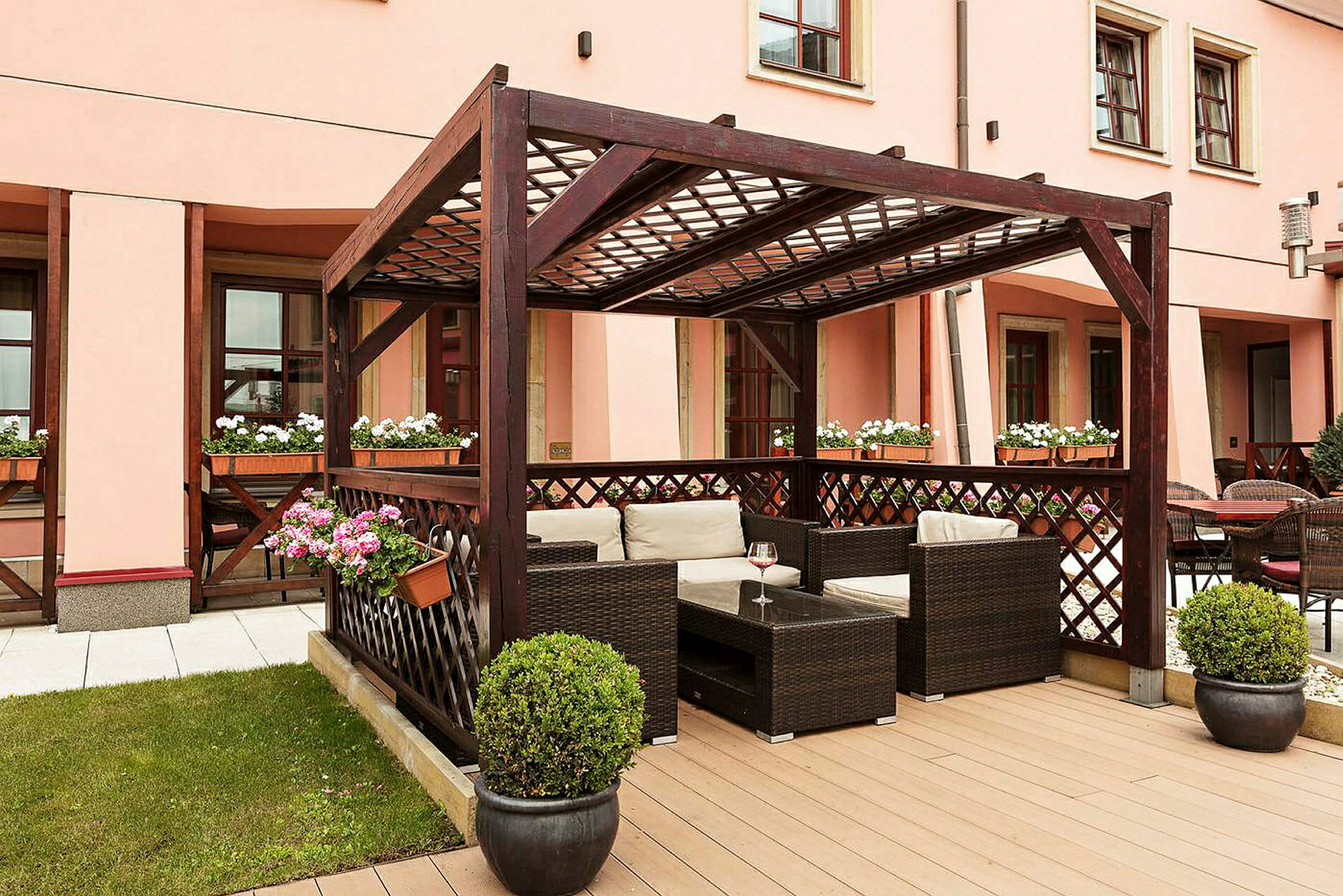 Bildet viser en uteplass med en pergola i mørkt tre, møblert med rottingstoler og bord. Blomsterpotter med fargerike blomster pynter området. (AI-generert tekst)