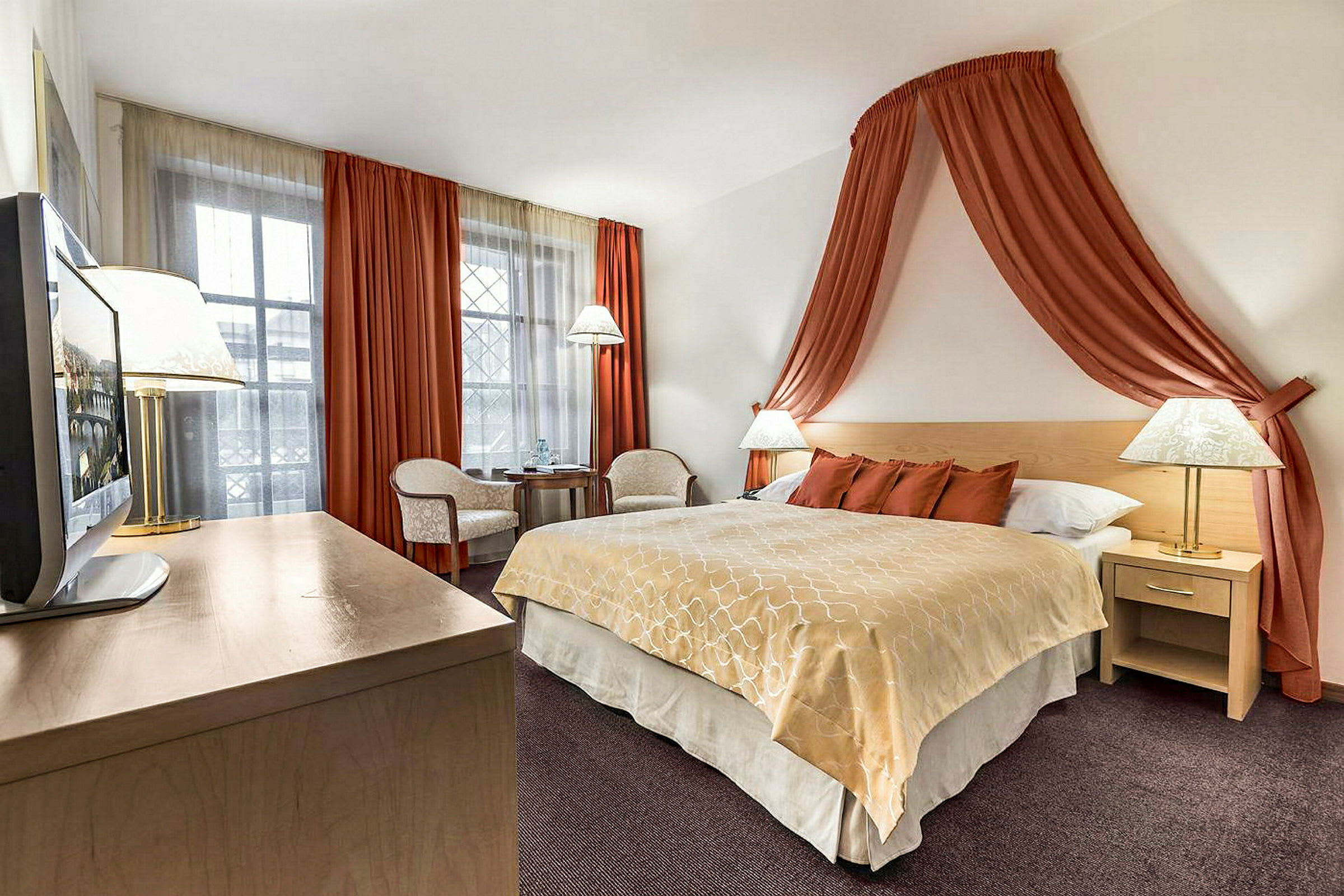 Et hotellrom med en stor seng dekket med beige og oransje sengetøy. Sengen er omgitt av nattbord med lamper og har oransje gardiner hengende over som draperi. I hjørnet står et lite bord med to stoler foran et vindu dekket med hvite og oransje gardiner. (AI-generert tekst)
