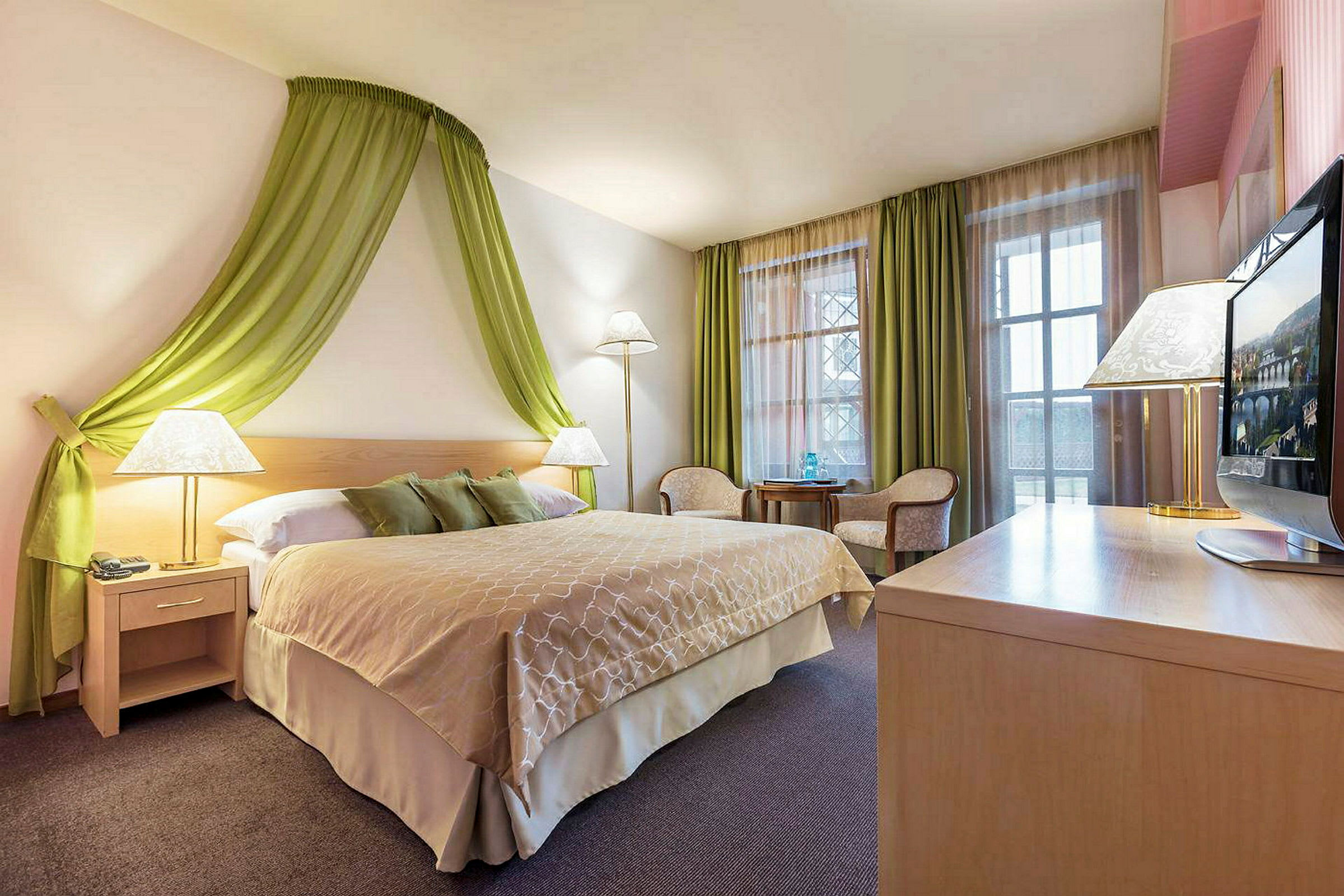 Et hotellrom med en dobbeltseng, grønne gardiner og veggdekorasjoner, sittemøbler og et skrivebord med en TV. (AI-generert tekst)