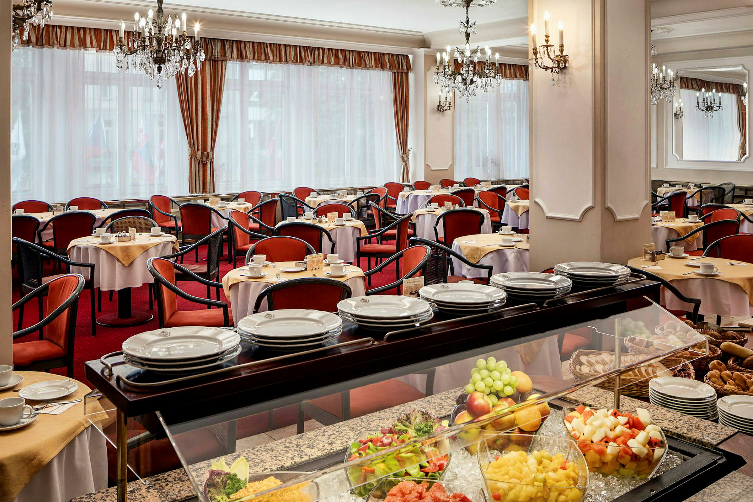 Spisesal med bord dekket med hvite duker og røde stoler. En buffet med frukt og brød er synlig i forgrunnen. (AI-generert tekst)