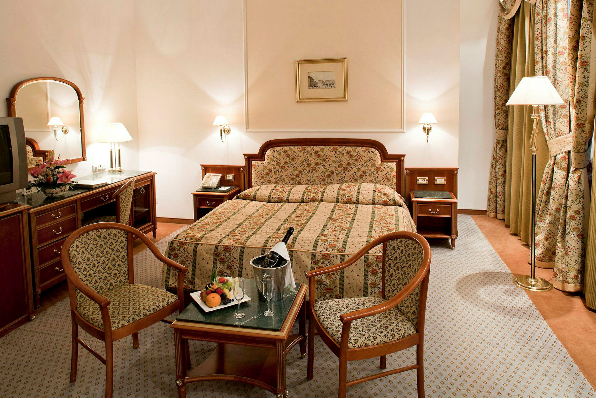 Hotellrom med dobbeltseng, to stoler, bord med frukt og champagne samt klassisk innredning. (AI-generert tekst)